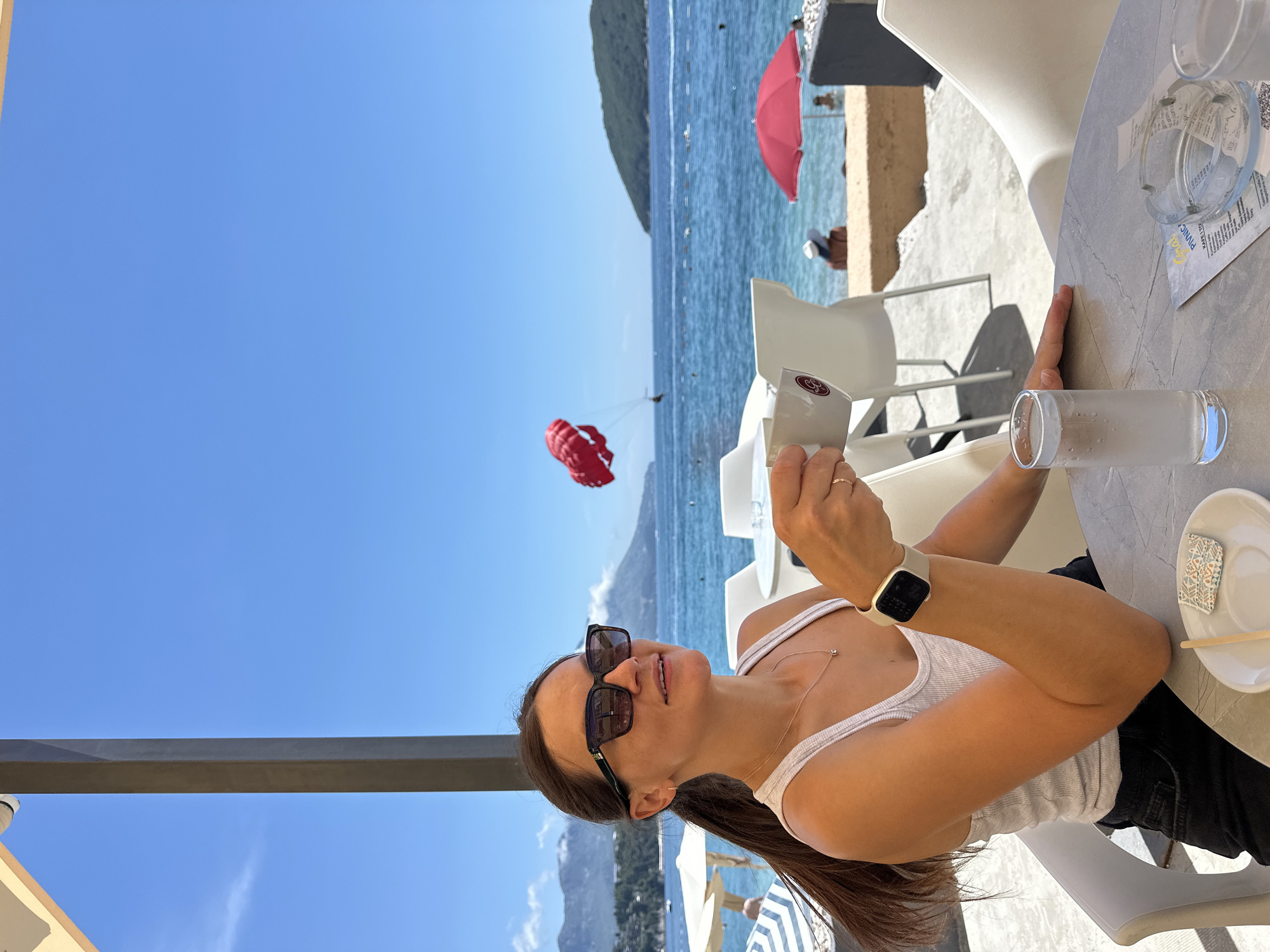 A woman is sitting at a table outdoors, holding a mug and looking up with a smile. She is wearing a white tank top, black pants, and sunglasses. A smartwatch is visible on her left wrist. In the background, the sea and mountains are visible, with a red parasail in the air and a red umbrella on the beach. The scene suggests a sunny day at a beachside cafe in Budva, Montenegro. The water appears calm, and the sky is clear blue, indicating pleasant weather. The woman's expression conveys happiness and relaxation. A glass of water and a plate with chopsticks are on the table. There is some visible text on a menu or paper on the table, but it is not legible.