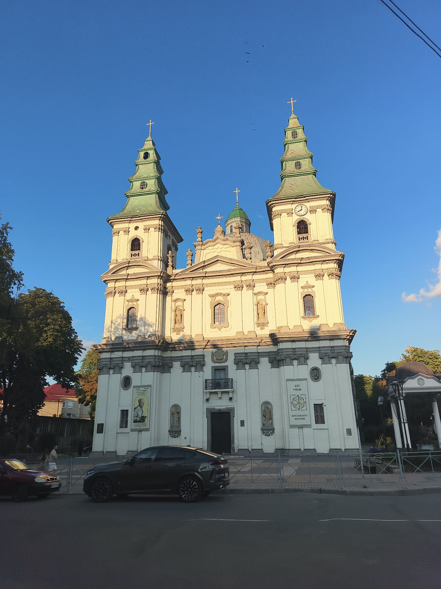The image captures a frontal view of a prominent church with two tall, green-capped bell towers flanking a central facade. The church is a light color, possibly white or cream, and features ornate architectural details, including columns, statues, and window frames. A clock face is visible on the right-hand bell tower. The sky is a clear, vibrant blue. In the foreground, a dark-colored SUV is parked on the street in front of the church. To the left, a red sedan is also parked. A sidewalk with a decorative metal fence separates the street from the church. Trees with green and autumnal foliage are visible on the left and right sides of the church, adding to the scenery. The scene is bathed in the warm light of what appears to be late afternoon or early evening. The church is the Church of the Virgin Mary's Nativity in Ternopil, Ukraine.