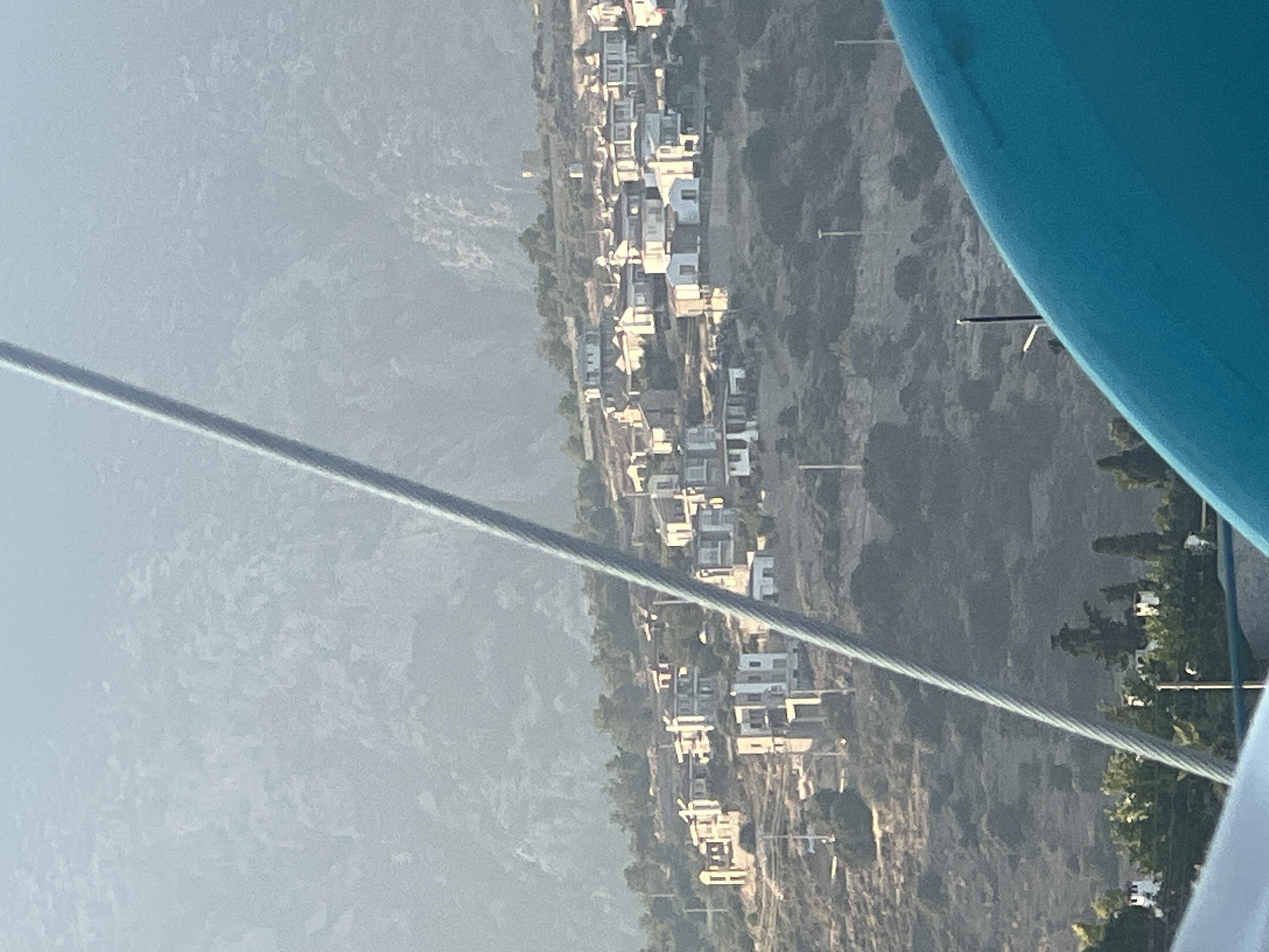 The image shows a hillside village with white and beige buildings clustered together. The buildings have red-tiled roofs and some have balconies. The hillside is covered in dry vegetation and scattered trees. A large grey cable runs diagonally across the image, obscuring parts of the scene. In the foreground, a bright blue curved object occupies the bottom right corner. The background consists of a large, hazy mountain. The lighting suggests it is either early morning or late afternoon.