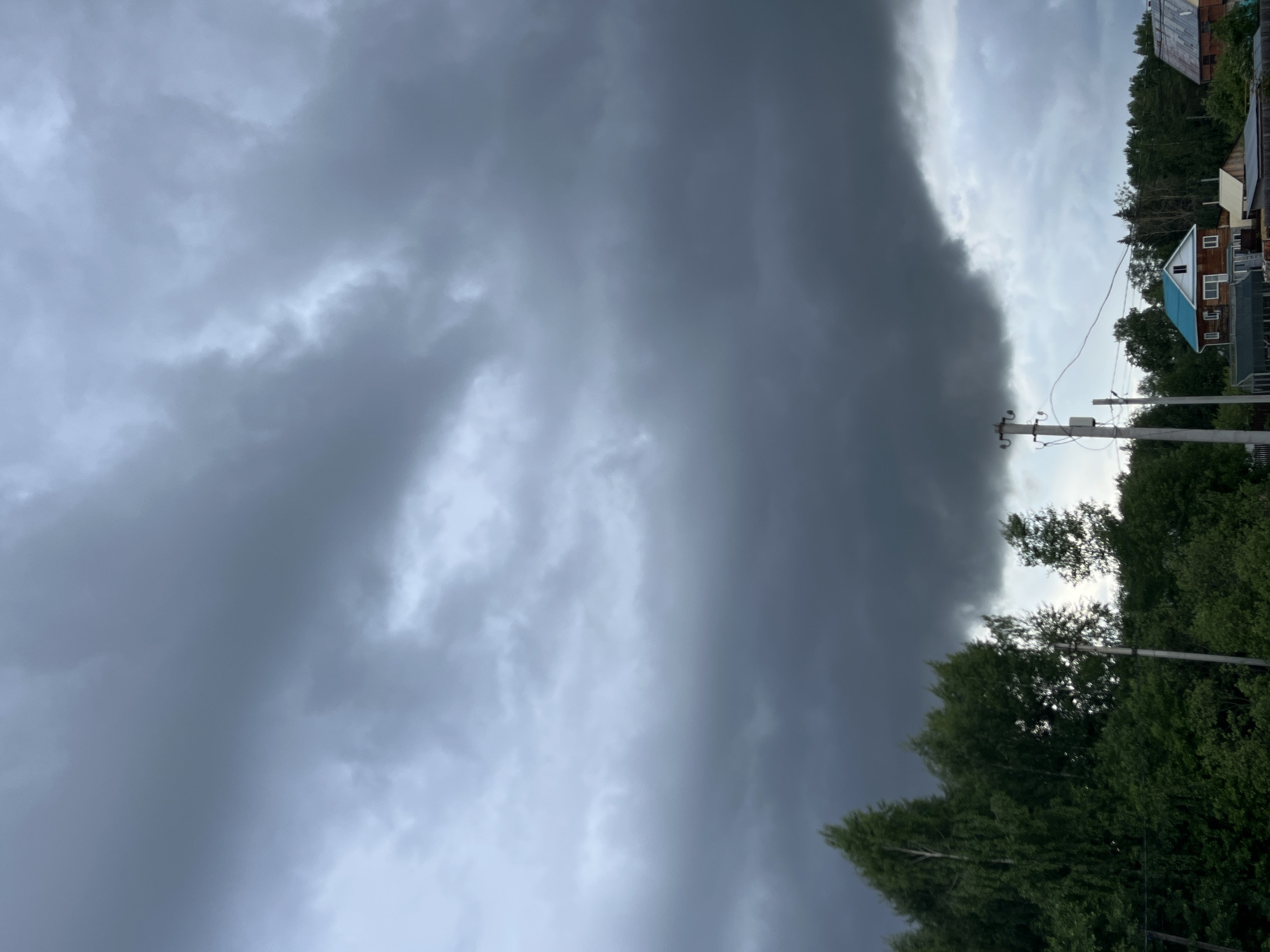 The image captures a dramatic sky over a rural or suburban landscape in Chaikovsky, Russia. A striking weather phenomenon is depicted, with the sky sharply divided into two distinct halves. The left side is dominated by a dense, dark gray to almost black cloud bank, characteristic of an approaching or intense storm. This heavy cloud formation creates a powerful, ominous presence.
In contrast, the right side of the sky features lighter gray clouds, which, while still overcast, appear less threatening and brighter, suggesting the storm front is either yet to fully arrive or is receding.
Below the sky, a green tree line stretches across the landscape, with dense foliage appearing verdant. Amidst the trees, several utility poles with power lines are visible, indicating an inhabited area. Portions of multiple houses can be seen peeking through the trees, one of which has a distinctive bright blue roof, adding a subtle splash of color to the scene.
The image conveys the raw power of nature and the immediate impact of a significant weather event. The time of day is not precisely discernible due to the heavy cloud cover, but it appears to be daytime. There are no visible people or text in the image.
