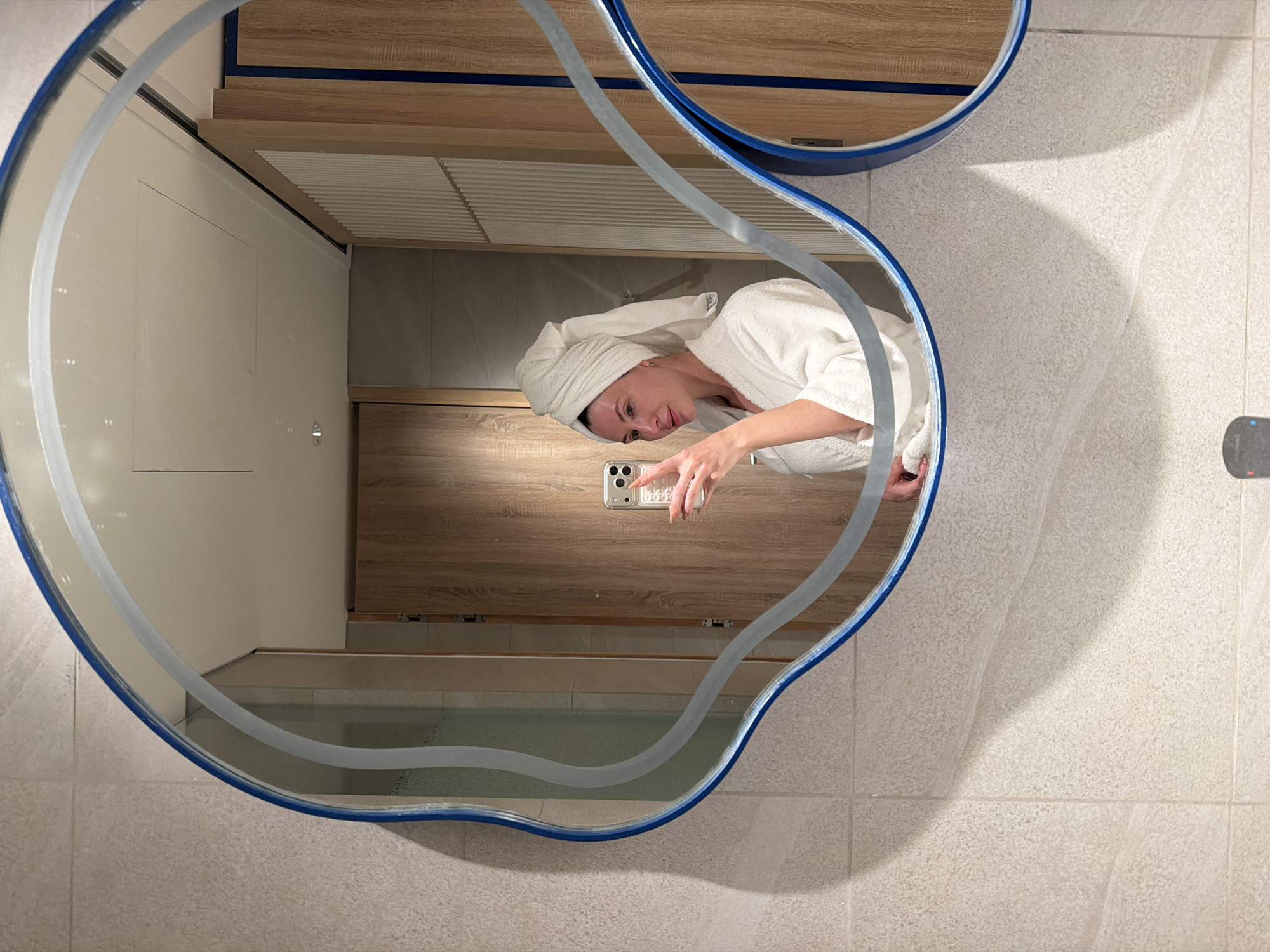 A woman is depicted taking a mirror selfie in a bathroom or hotel room located in Dong Ninh Hoa Ward, Vietnam. She is Caucasian, wearing a white terry cloth bathrobe, and has a white towel wrapped around her head in a turban style. Her left hand, adorned with light-colored manicured nails, holds a silver-colored smartphone with a visible triple-lens camera array and a textured, clear phone case. She is looking towards the phone's reflection in the mirror. The mirror itself is irregularly shaped, featuring a unique wavy outline with a prominent blue border on the outermost edge and an inner clear, wavy line. The room's interior, reflected in the mirror and visible around it, consists of light-colored, textured square wall tiles, light wood-grained cabinetry or doors, and a ceiling with recessed lighting. Some wooden elements feature vertical slats and a dark blue or black vertical accent strip. A partial view of a sink fixture or control panel is visible at the bottom edge of the frame.