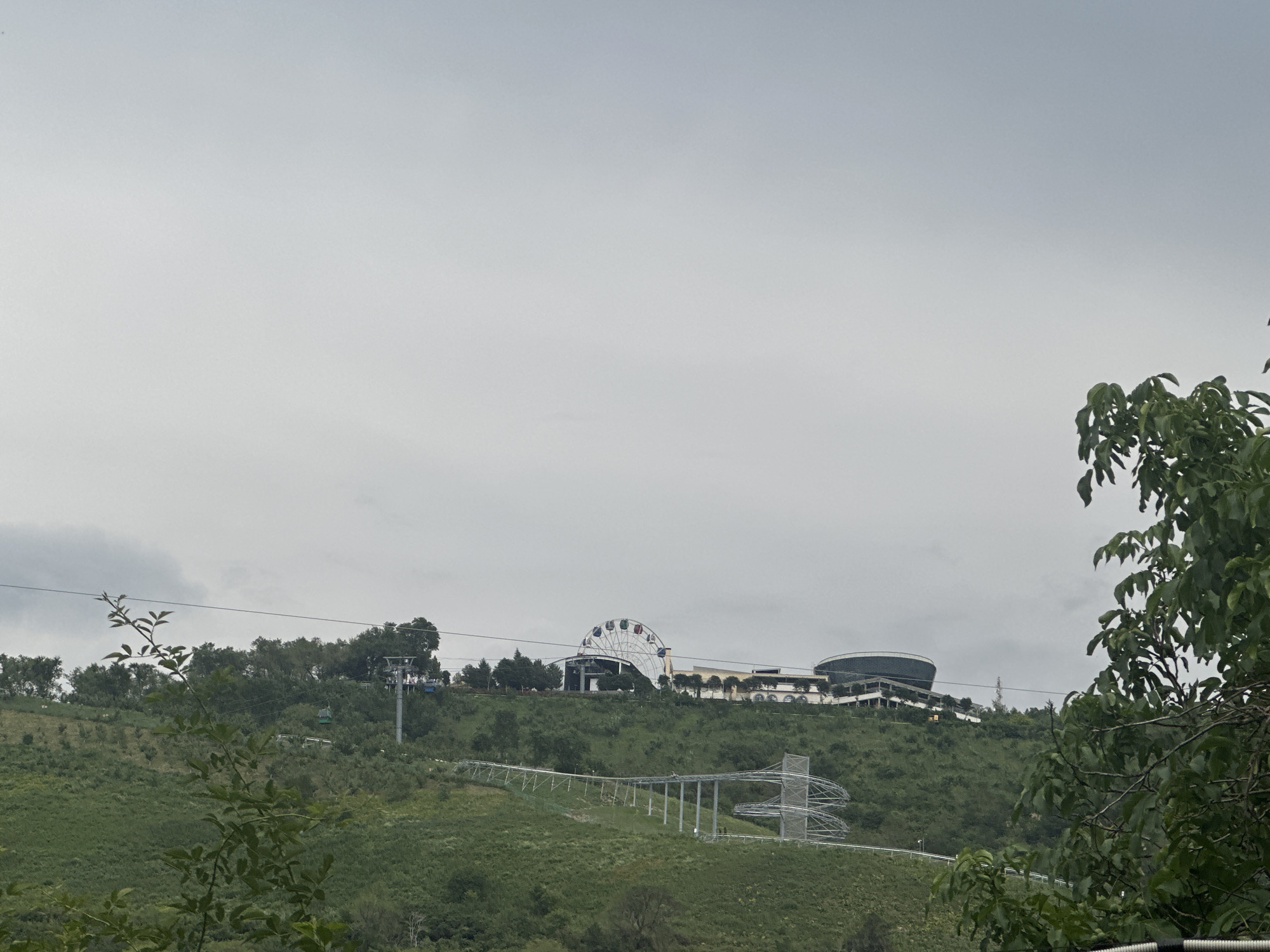 The image depicts a scenic view of Kok Tobe hill in Almaty, Kazakhstan, under an overcast sky. The foreground is dominated by lush green foliage, partially obscuring the view of the hill. In the mid-ground, the green slopes of Kok Tobe are dotted with trees and shrubs. A prominent feature on the hill is a roller coaster track, winding down the slope with its metallic structure visible against the green backdrop. Further up the hill, a Ferris wheel stands out, its colorful cabins contrasting with the subdued lighting. Adjacent to the Ferris wheel are modern buildings, likely part of an entertainment complex, with a large, circular building being particularly noticeable. Several gondola cabins are also seen traversing a cableway, ascending the hill. The overall scene suggests a recreational area with various attractions for visitors.
