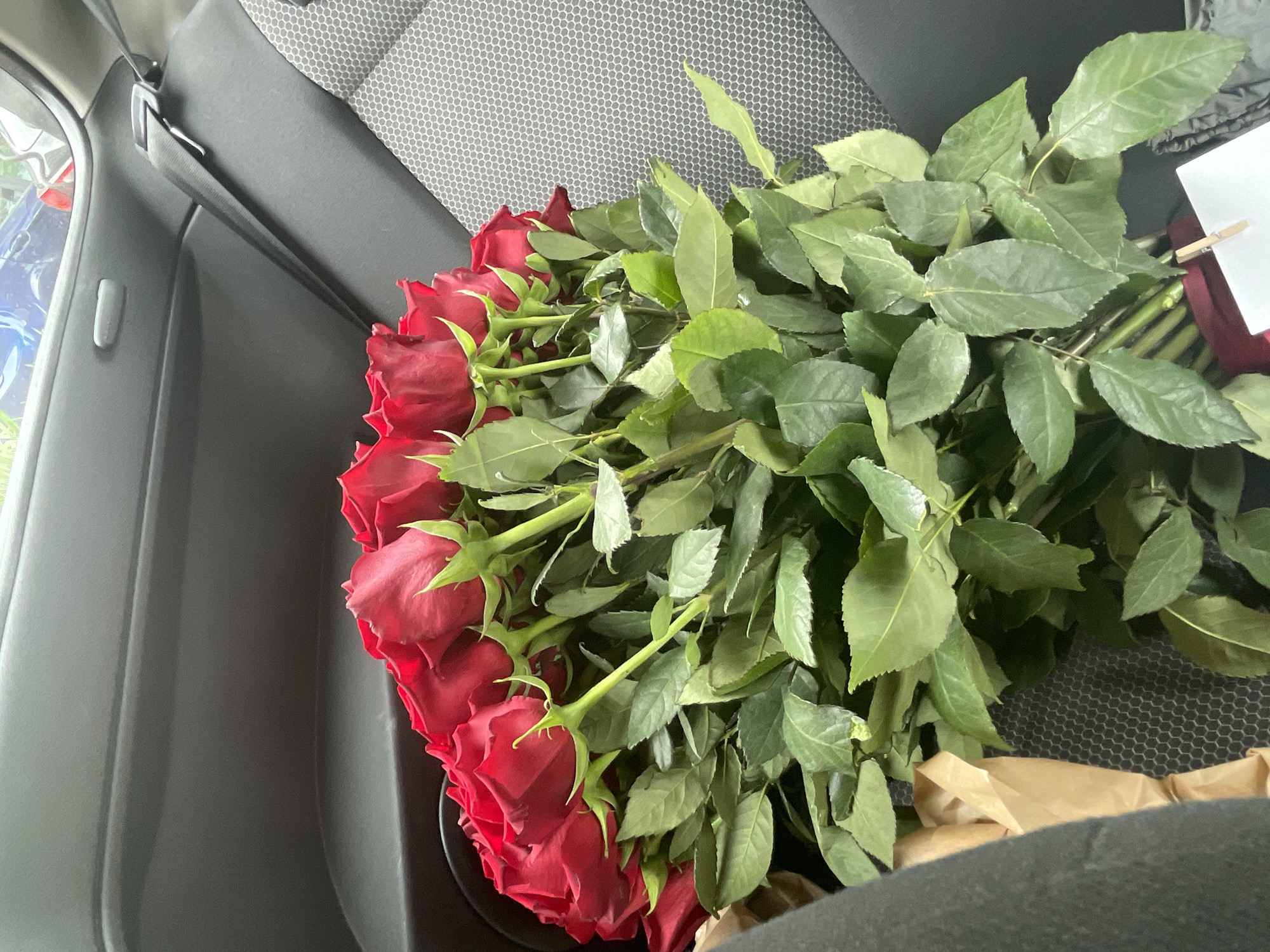 A bouquet of red roses with green leaves lies on a gray seat, likely inside a vehicle, as evidenced by the seat texture and the car door visible in the image. The location is unknown. The roses appear freshly cut and are arranged in a bunch, suggesting they were recently purchased or gifted. A small, white card is attached to the bouquet.