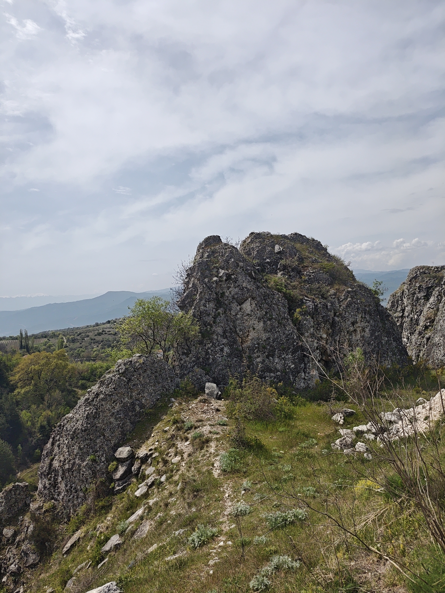 This media file captures a rugged, natural landscape under an overcast sky. The scene is dominated by large, craggy rock formations in the midground and foreground, interspersed with patches of green grass, small trees, and dry bushes.
In the immediate foreground, a rough, earthy path-like area is visible, covered with sparse green and yellowed grass and scattered rocks. The ground slopes upwards towards prominent, dark grey rock outcrops that show distinct textures and crevices. A small, bright green deciduous tree grows out of one of these central rock formations, with further dense green foliage and trees visible in a valley or lower terrain to the left. More distant hills and mountains, rendered in soft blue and grey tones, stretch across the horizon in the background, suggesting significant depth and distance.
The sky above is filled with soft, diffused white and light grey clouds, indicating an overcast or partly cloudy day. No specific activity is occurring, and no people or animals are visible. The time of day is daytime, with ample natural light. The overall impression is one of a wild, untouched environment, possibly a mountainous or hilly region with a temperate climate. There is no visible text in the image.