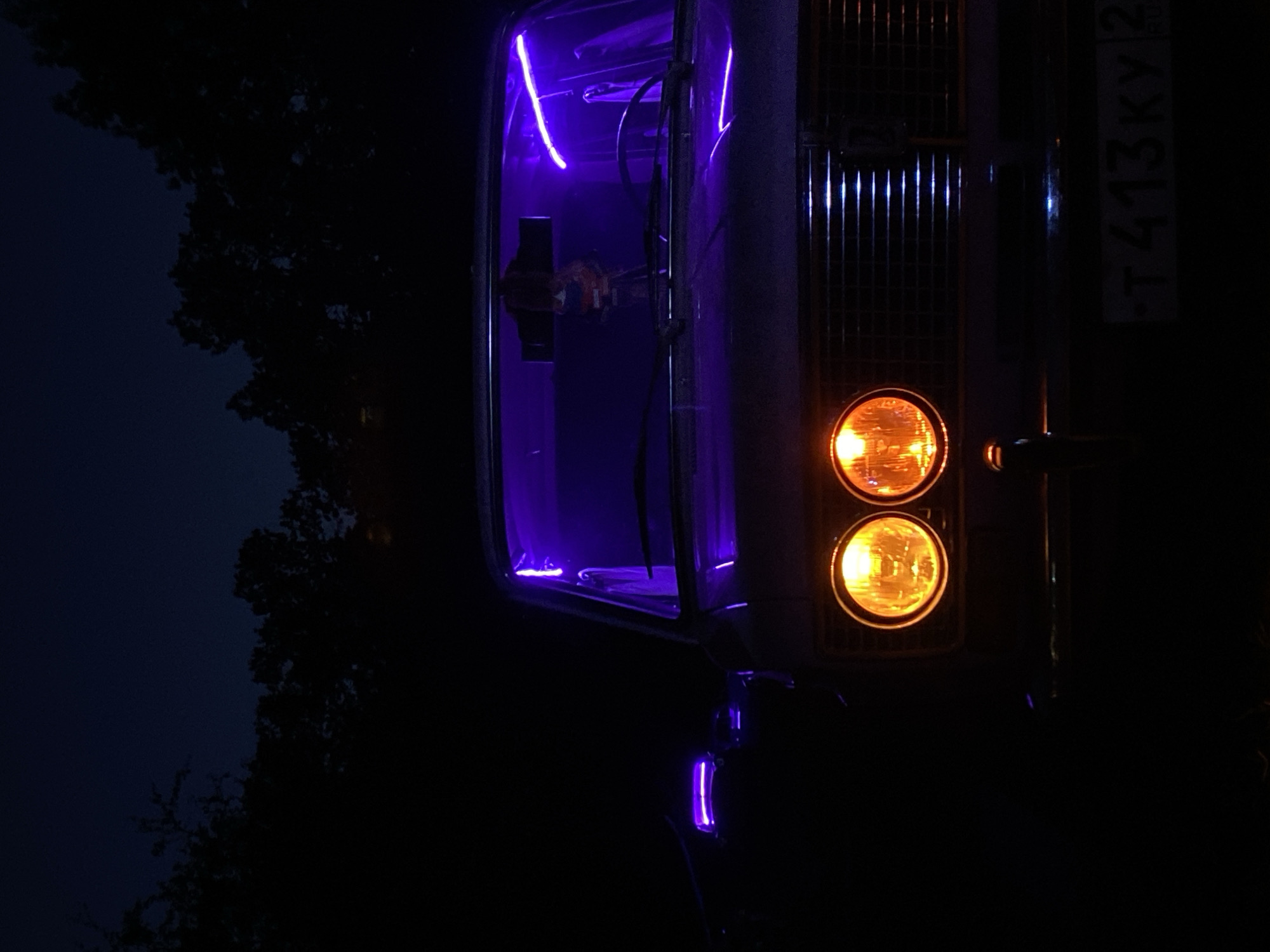 A close-up, low-angle shot depicts the front of a dark-colored vintage car parked at night in Bezymyannoye, Russia. The car's dual round headlights are illuminated with a warm orange glow. A dark grille is visible between the headlights. Purple LED strip lights are installed along the windshield's interior and on the driver's side mirror, casting a vibrant violet hue. The dark sky is visible above, with silhouetted trees forming a dark backdrop. A license plate reading "T 413KY 2" is affixed to the front bumper. No people are present in the frame.