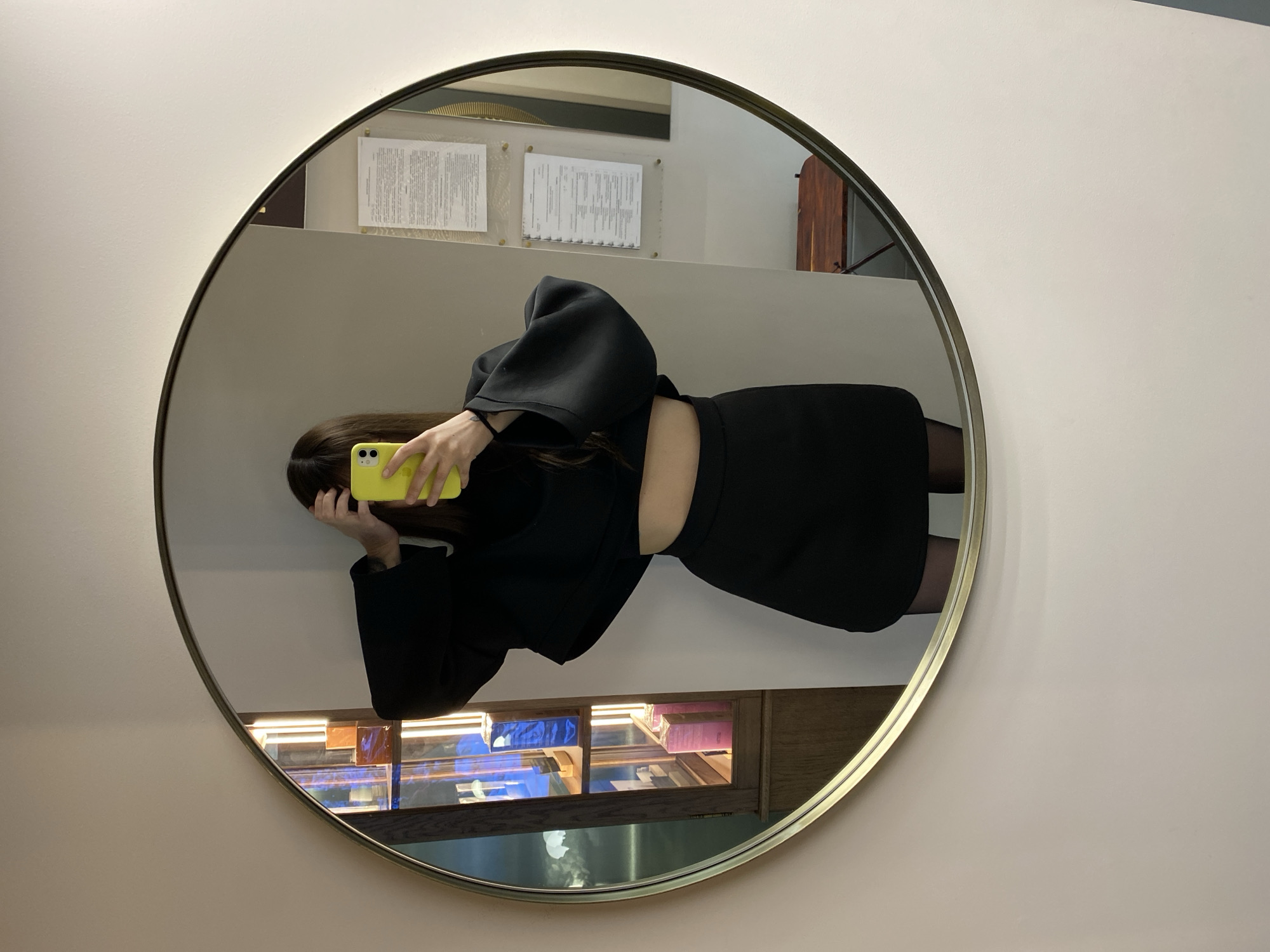 An image, oriented sideways, depicts a person taking a mirror selfie in a round, gold-framed mirror. The person is holding a vibrant yellow smartphone, which obscures their face, but shows their long dark hair and a hand with a subtle wrist tattoo. They are dressed in a black long-sleeved top with wide, voluminous sleeves and a black skirt or pair of shorts, revealing a sliver of bare midriff. Dark hosiery or leggings are visible on their legs.
The mirror is set against a plain, light-colored wall. Within the reflection, behind the person, two sheets of paper with indistinct text are affixed to a wall or surface. Below these documents, a wooden display cabinet with glass shelving is visible, illuminated from within and showcasing various products, some casting a blue light. Part of a dark wooden structure, possibly a door or wall panel, is also reflected. The overall setting appears to be an indoor commercial environment, like a retail store or salon, in Votkinsk, Russia. No specific text on the documents is legible. The time of day and weather are not discernible.