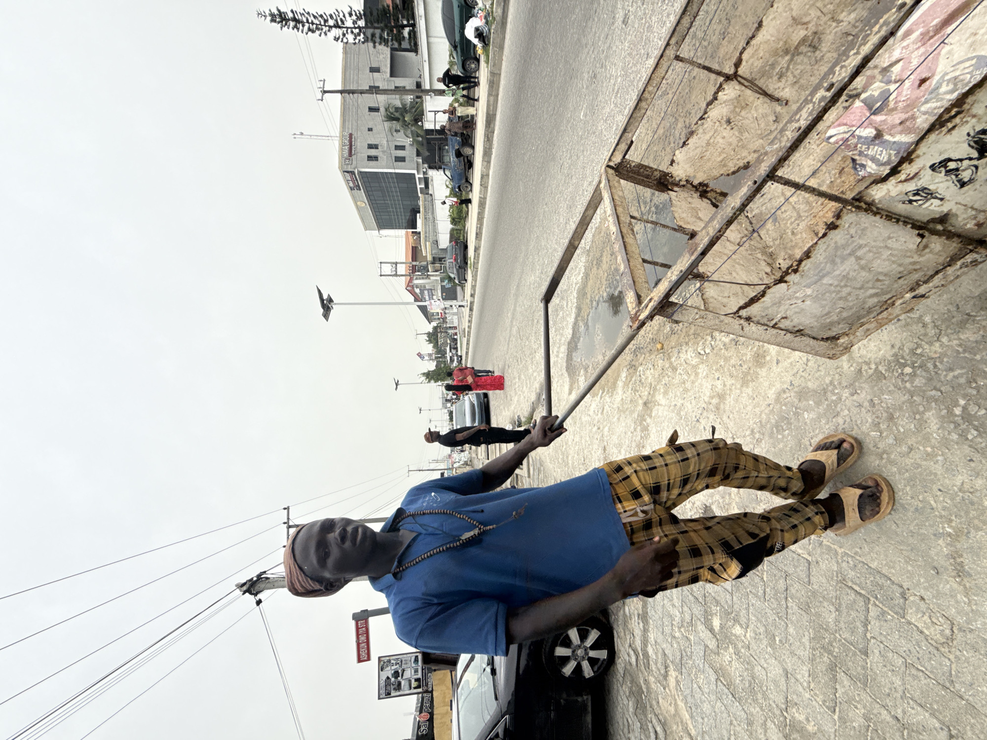 A man is standing on a sidewalk in Ado, Nigeria. He is pulling a dirty, wheeled cart with a metal frame and weathered wooden panels. The man is wearing a blue short-sleeved polo shirt, plaid pants, and sandals. He has a beaded necklace and a cap on his head. Behind him is a car parked on the side of the road. In the background is a street with buildings, pedestrians, and other vehicles. A street sign reading "ASHEKEWUN OWO OTU STR" is visible. The sky is overcast.
