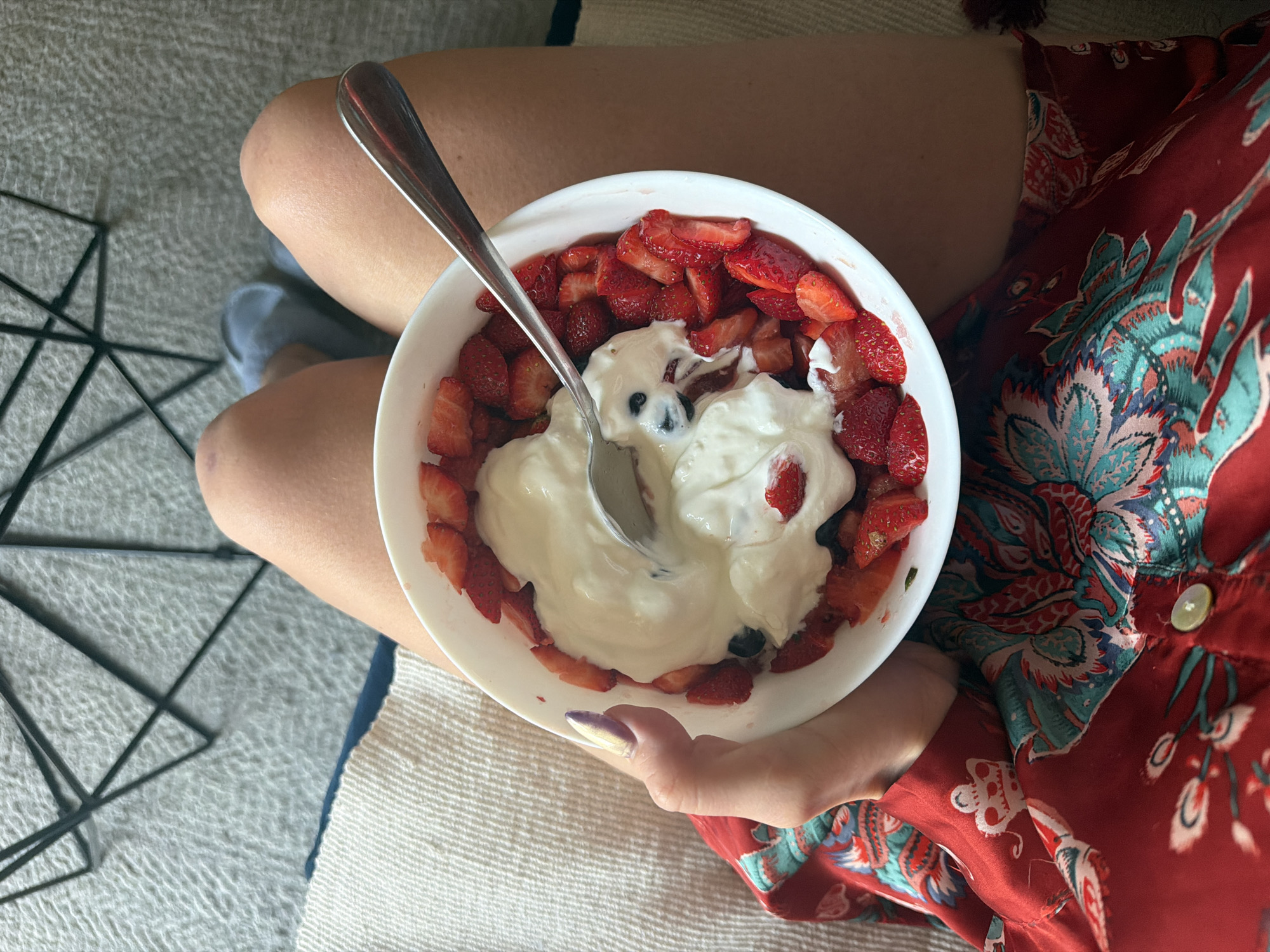 A top-down, eye-level shot shows a person holding a bowl of food while sitting on what appears to be a sofa or cushioned area.
The bowl, which is white, contains cut strawberries around the edge and a dollop of white yogurt or cream in the center. A silver spoon is plunged into the yogurt.
The person holding the bowl has light skin and painted fingernails, with one hand visible. They are wearing a red shirt or dress with a pattern that includes teal, white, and red elements.
The seating area has a light-colored, textured surface. There is also a dark geometric metal object nearby.
The setting appears to be indoors. The location, Belgrade, Serbia, is not directly identifiable within the image.