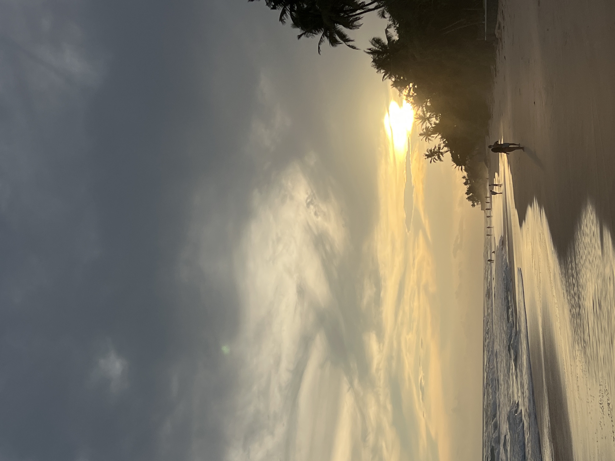 The image captures a tranquil beach scene in Habaraduwa, Sri Lanka, during the golden hour of either sunrise or sunset. The sky, occupying the upper portion, transitions from a dark, cloudy gray on the left to a brilliant golden and orange hue towards the right, where the sun is low on the horizon. The bright sun, partially obscured by clouds and foliage, radiates a strong glow.
Silhouetted palm trees line the right edge of the frame, their dark forms contrasting sharply with the bright sky and sun. Below, a wide expanse of sandy beach stretches across the lower portion of the image. The wet sand near the water's edge is highly reflective, mirroring the warm colors of the sky. Gentle waves can be seen along the shoreline.
Several individuals are present on the beach. In the mid-ground, a prominent figure stands facing the ocean, their silhouette defined against the illuminated sand. Further in the distance, closer to the water, two other smaller figures are also visible, engaged in a leisurely stroll. The overall scene evokes a sense of peace and natural beauty at this coastal location. No text is visible in the image.