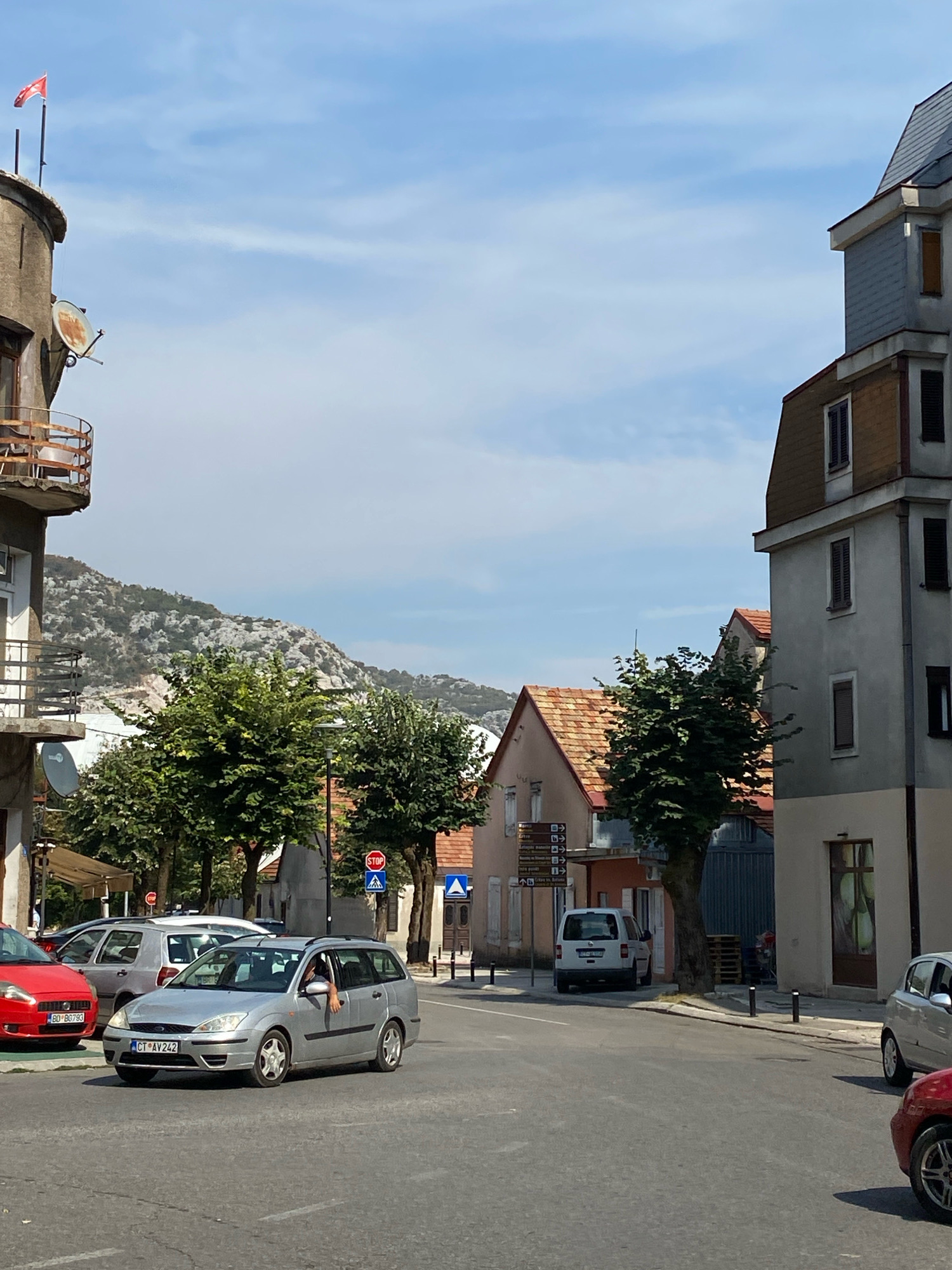 The image captures a street scene in Cetinje, Montenegro, on a bright, sunny day with a partly cloudy sky. In the foreground, a silver station wagon, identified by its license plate CT AV 242, is driving on the asphalt road. A person's arm is visible extending from the driver's side window. To the left, a red hatchback with license plate BD BO793 is parked. Behind the silver car, another silver station wagon is visible.
The environment features buildings with tiled roofs and modern facades, some adorned with shutters. A tall, narrow building with a distinct mansard roof is on the right side of the frame. Trees with lush green foliage line the street, and a rocky, mountainous landscape is visible in the background. Street signs, including a stop sign, are present, indicating traffic control. Satellite dishes are mounted on the side of a building on the left, and a flag flies from a pole on the same structure. Pallets are stacked near a building entrance on the right.