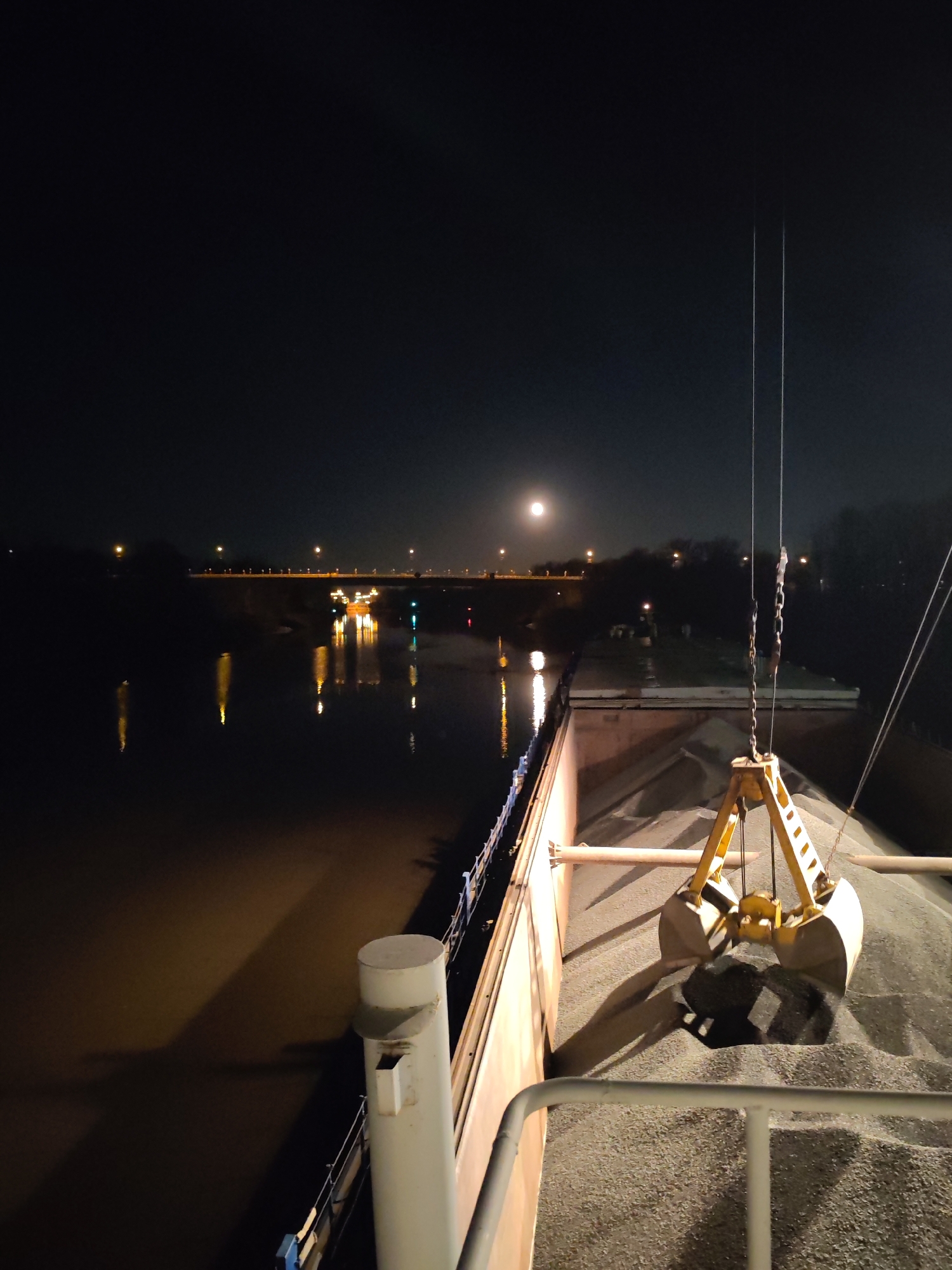 The image depicts a nighttime scene, likely in or near Yakhroma, Russia, judging by the reference to the city. The foreground focuses on the deck of a barge or similar vessel, filled with what appears to be gravel or aggregate material. A yellow, clam-shell type crane is positioned above the material, ready for operation. The crane is suspended by cables and chains. The deck of the barge is illuminated, possibly by artificial lighting. In the background, across a body of water (likely the Yakhroma river or a canal), a bridge is visible, illuminated by streetlights which reflect in the water. The moon is visible in the night sky. The lighting suggests the scene is captured at night or during very low-light conditions. There are no people visible in the image.