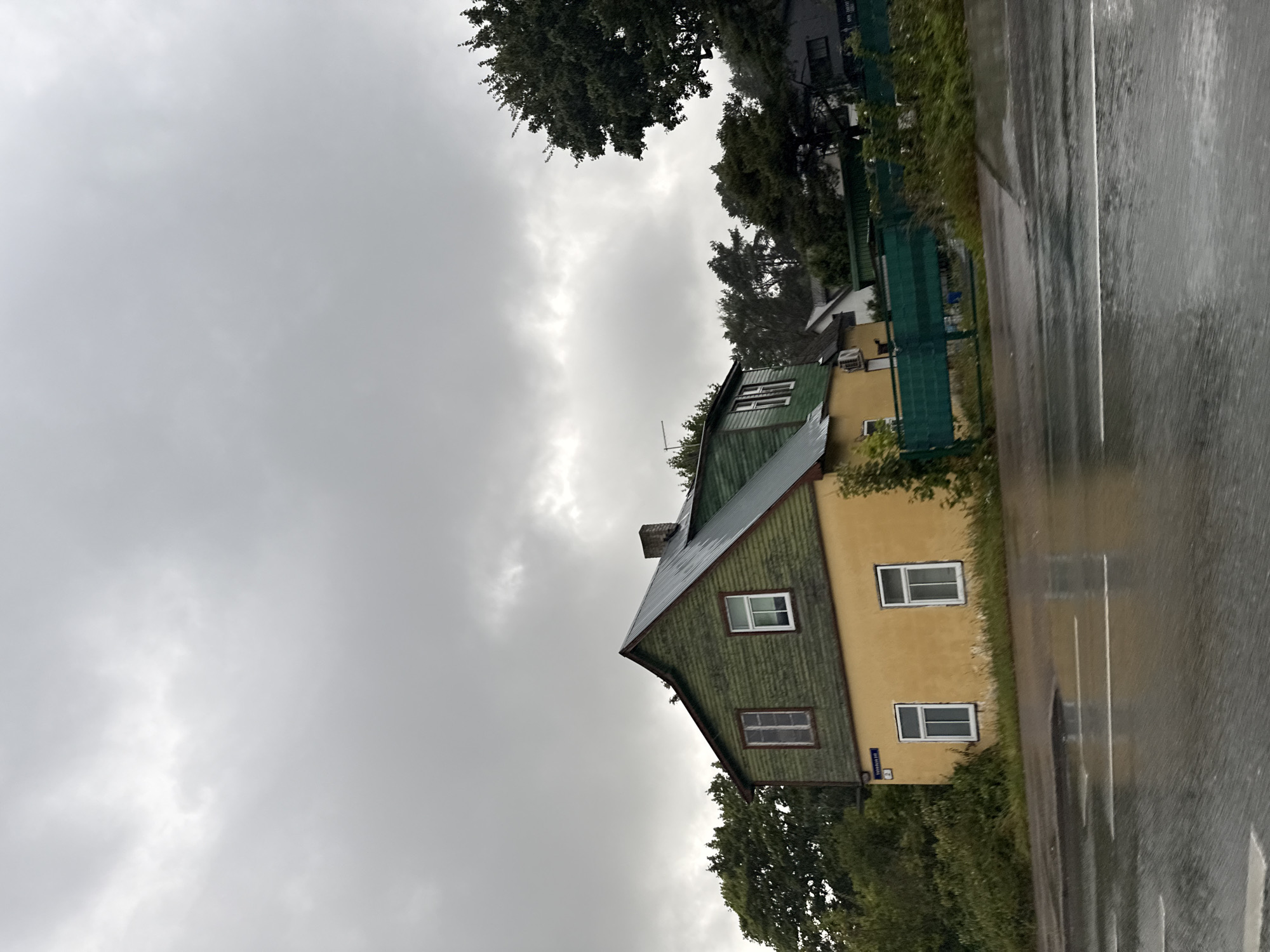 The image shows a weathered, two-story house with yellow lower walls and green upper walls, situated in Tallinn, Estonia, on a cloudy day. The house has a dark green, sloped roof with a visible chimney. Several windows with white frames are present on both floors. A street runs in front of the house, with white lane markings and reflections of the cloudy sky and surrounding vegetation on the wet pavement, indicating recent rain. A green fence runs along the property line, bordering the street. Lush green trees and bushes surround the house. In the background, other buildings and trees are visible under the overcast sky. A street sign with the number "2" and text in a foreign language is affixed to the corner of the house.