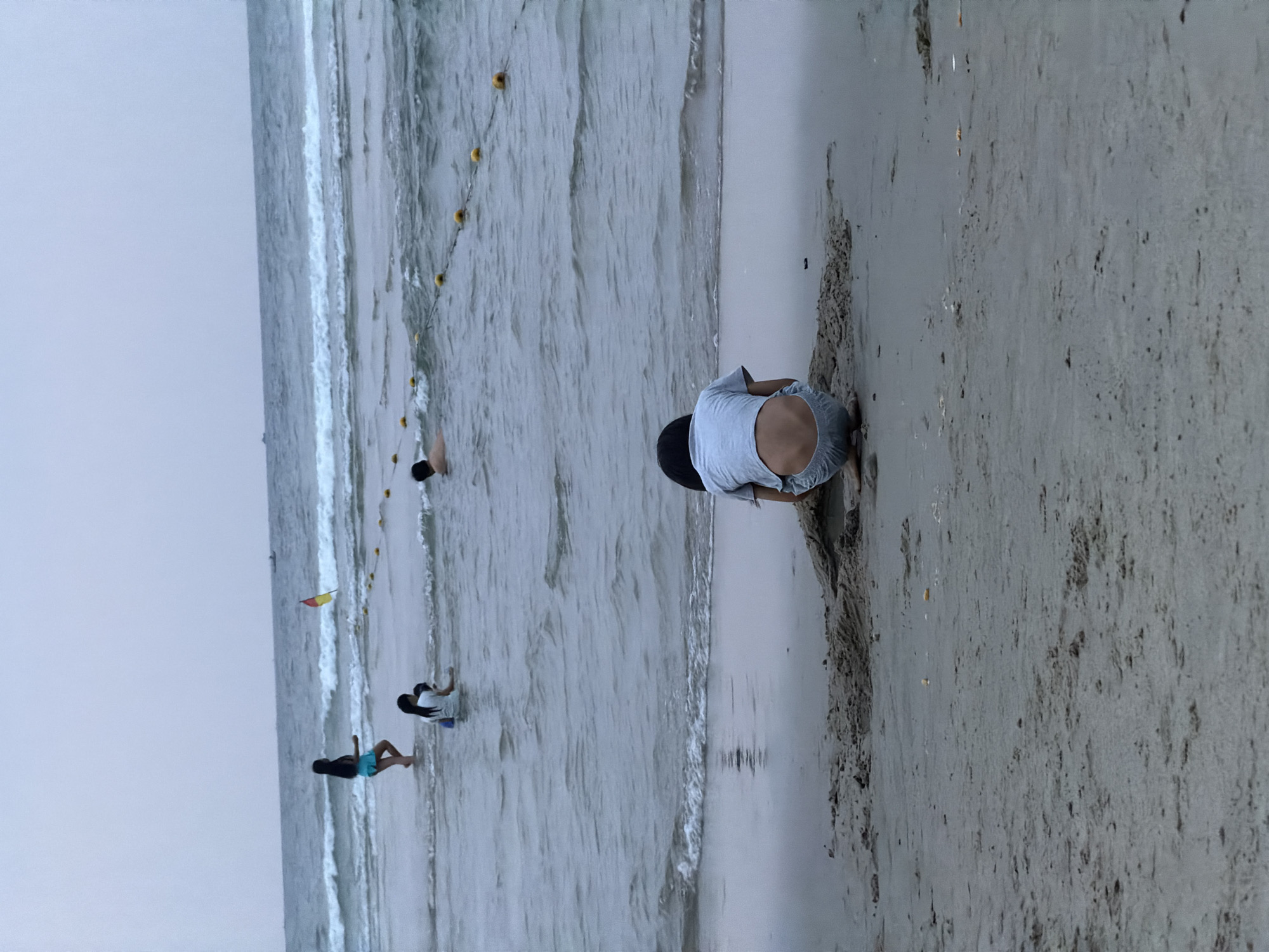 The image depicts a beach scene with several people interacting with the water and sand.
A person is kneeling and digging in the sand on the right side of the image. In the water, there is a line of yellow buoys marking off a swimming area. Further into the water, a person is seen floating. Closer to the viewer and in the water, there is another person. Another individual stands at the edge of the water near the left edge.
The environment consists of a sandy beach and ocean under an overcast sky. The scene appears to be in Đà Nẵng, Vietnam.