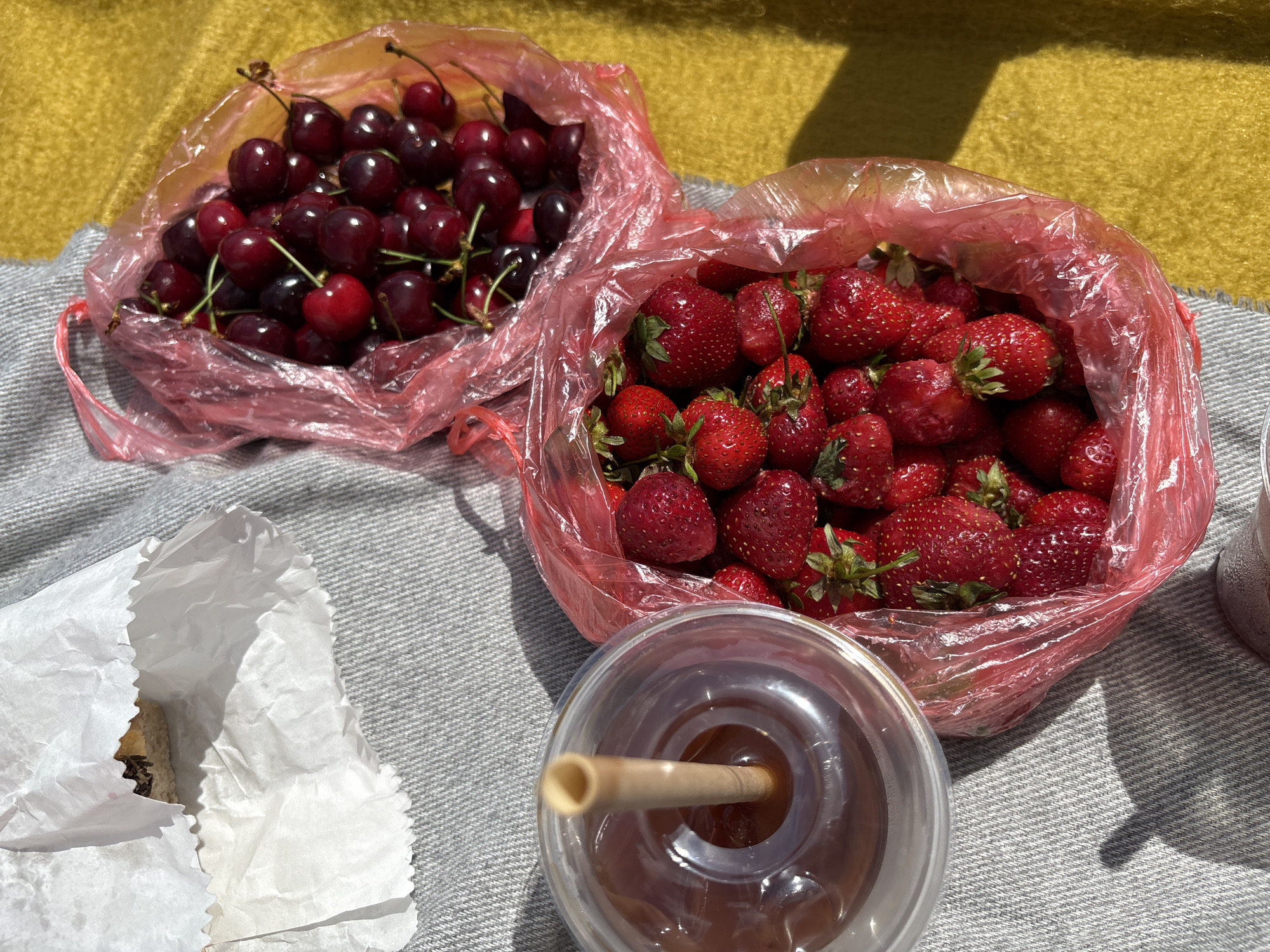 The image shows a picnic scene, likely in an outdoor location in Vracar Urban Municipality, Serbia. There are two bags of fruit: one filled with cherries and the other with strawberries. A clear plastic cup containing a dark beverage with a light-colored straw is visible near the strawberry bag. A white paper bag, possibly containing food, is also present. The items are placed on a gray and white striped surface, possibly a blanket or cloth, and a portion of a yellow surface is visible in the background. The lighting suggests it is daytime.