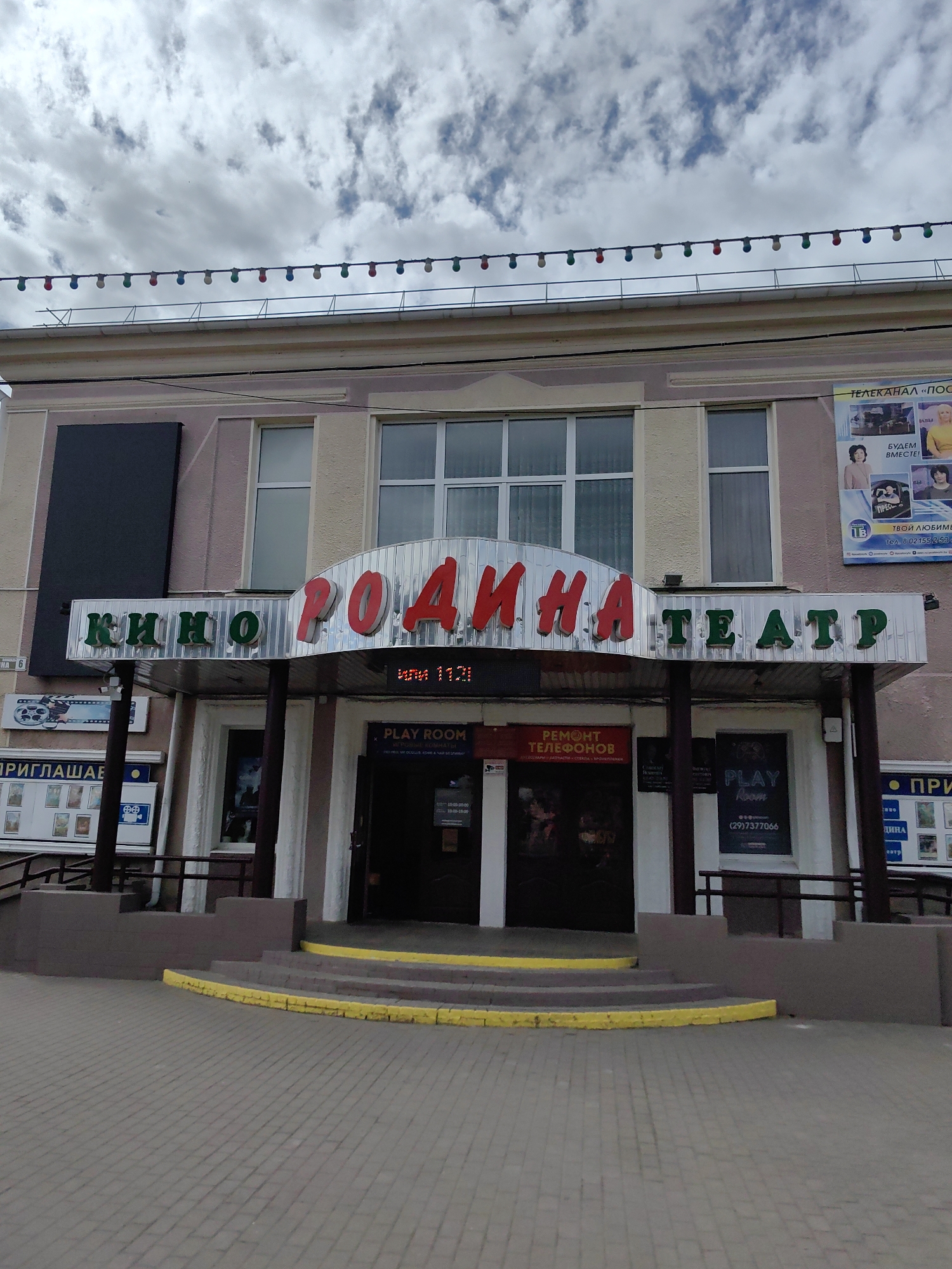 This is an exterior, eye-level, medium shot of the Rodina Cinema in Pastavy, Belarus. The cinema's facade is visible, with a prominent marquee displaying the name "КИНО РОДИНА ТЕАТР" in red and green lettering. Below the marquee, there are entrances with glass doors and posters. Signs indicating "PLAY ROOM" and "РЕМОНТ ТЕЛЕФОНОВ" (Phone Repair) are also visible. The building has a classical architectural style with large windows. A string of colorful lights is strung across the top of the building, and the sky above is partly cloudy. The foreground shows a paved walkway with a raised entrance leading to the cinema. The overall impression is that of an older, established entertainment venue.