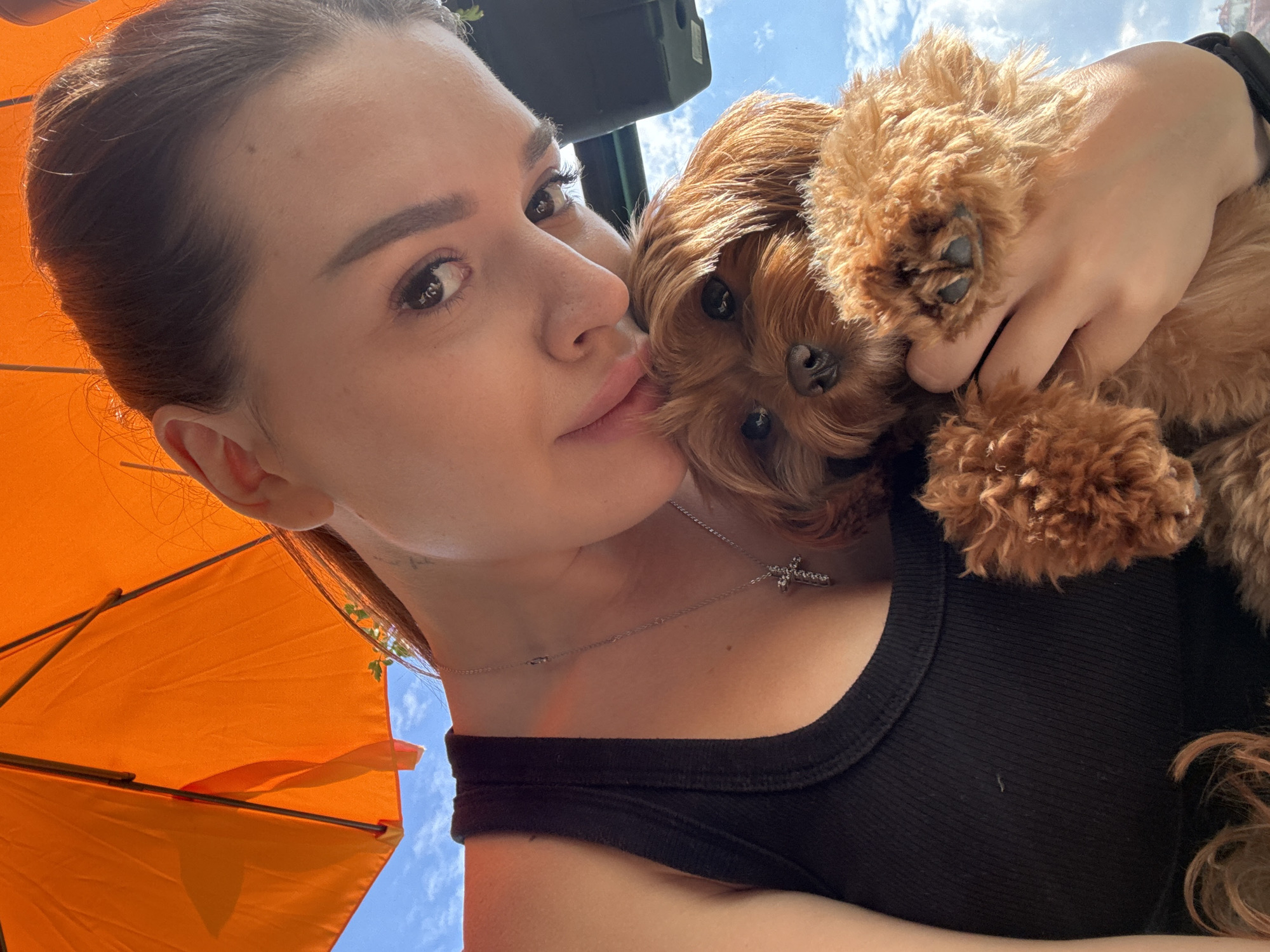In this close-up, high-angle shot, a woman with brown hair pulled back is holding a small, fluffy brown dog. She is wearing a black tank top and a silver cross necklace. The dog is nestled against her chest, with its paws resting on her hand. The background is dominated by an orange umbrella, suggesting an outdoor setting, possibly a cafe or park. The sky is visible behind the umbrella, with some clouds. The image does not provide specific landmarks or details that would allow for identification of a location in Prague, Czechia.