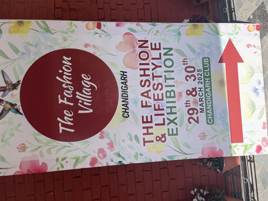The image shows a large banner advertising the "The Fashion Village" fashion and lifestyle exhibition, taking place on the 29th and 30th of March 2025 at the Chandigarh Club in Chandigarh, India (latitude: 30.764, longitude: 76.797). The banner is printed with a watercolor-style background of flowers and two hummingbirds in flight. The text is white, bold and in a sans serif font against a red circular background. The banner is attached to a brick wall. A red arrow at the bottom right corner points towards the direction of the event. There are flower pots on the ground, beside the banner.