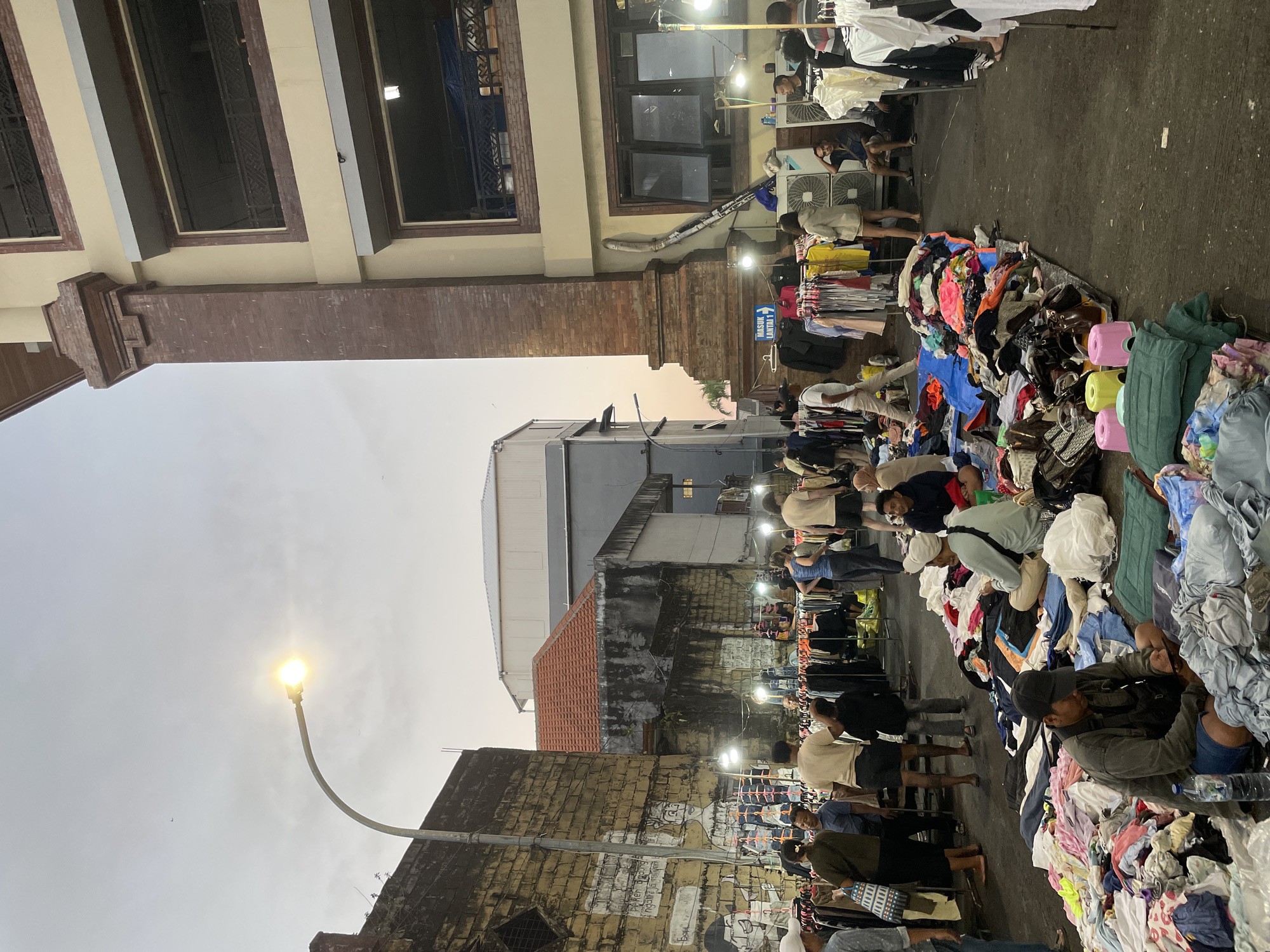 The image depicts a street market in Denpasar, Indonesia, likely a night market or an early evening street fair. The scene is characterized by an eclectic mix of goods for sale, primarily clothing, spread out on tarps and racks along a street. Several individuals are present, some browsing the merchandise, while others appear to be vendors or shoppers engaged in transactions. The environment is illuminated by streetlights and light bulbs strung overhead, casting a warm glow on the scene. The architecture visible includes a multi-story building with a distinctive brick facade on the right, and a weathered brick wall with graffiti on the left. The overall atmosphere suggests a bustling, casual marketplace.