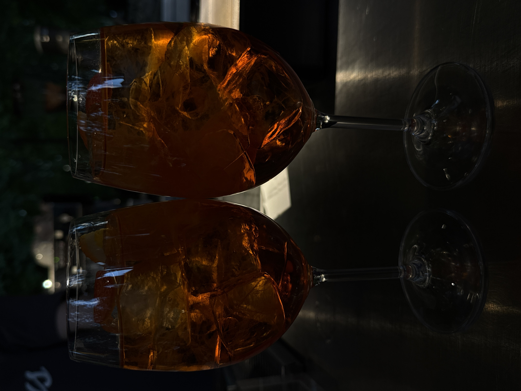 Two glasses filled with ice and an orange-colored liquid are positioned on a reflective surface. The glasses appear to be wine glasses. The scene is dimly lit. Based on clues in the background, the location is Yerevan, Armenia.