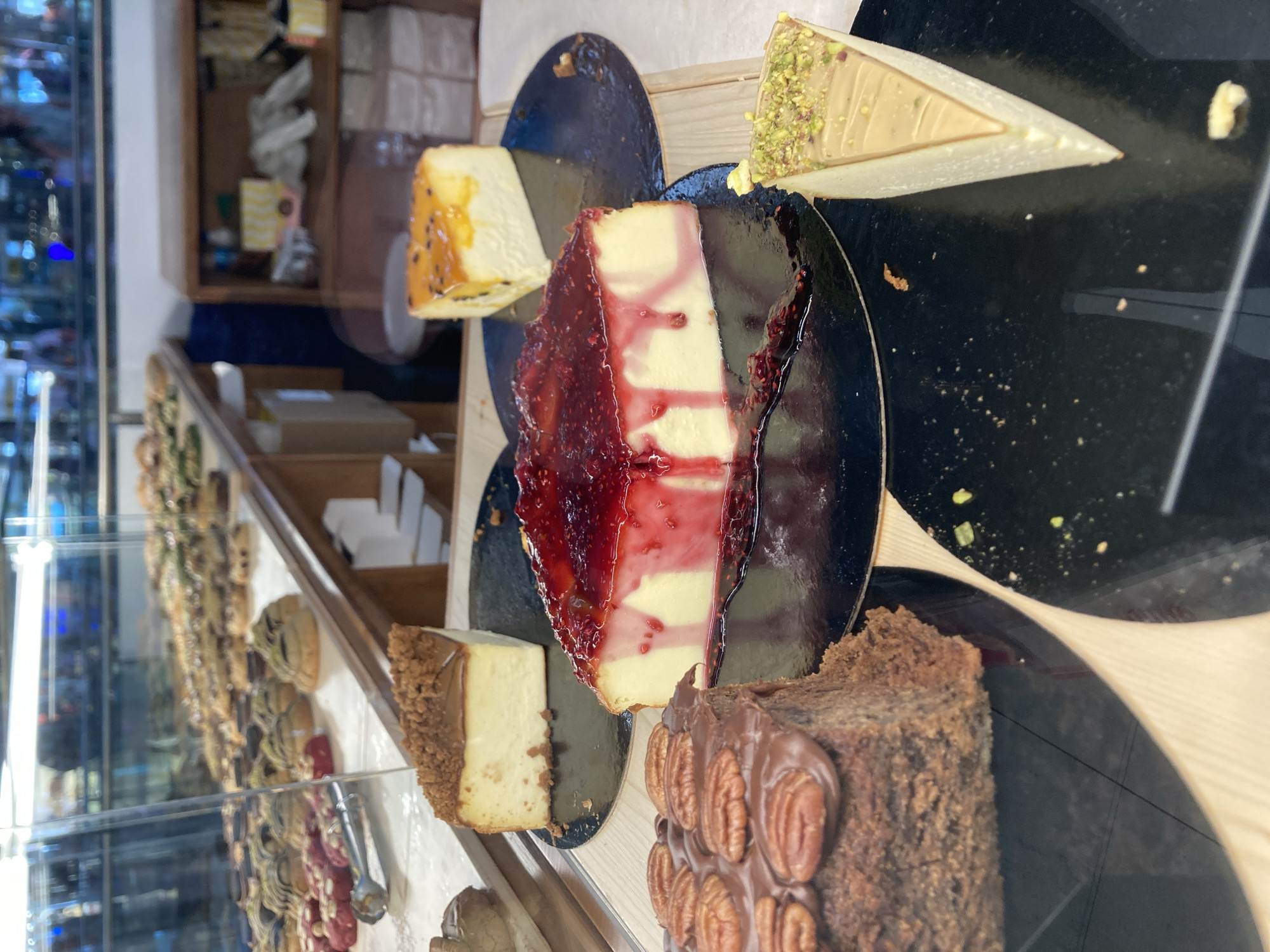 A display case in a Barcelona, Spain bakery holds an assortment of decadent desserts. The focus is on several slices of cheesecake. In the foreground, a rich chocolate cake topped with pecans and chocolate frosting sits to the left. To its right, a slice of cheesecake is generously covered in a bright red raspberry sauce. Further back, another cheesecake slice displays a vibrant orange topping, possibly passion fruit. To the right, a slice of cheesecake dusted with pistachios rests on a black circular plate. In the background, rows of smaller pastries are visible behind the glass, hinting at a wide selection of baked goods. The environment suggests a clean, well-lit commercial establishment.