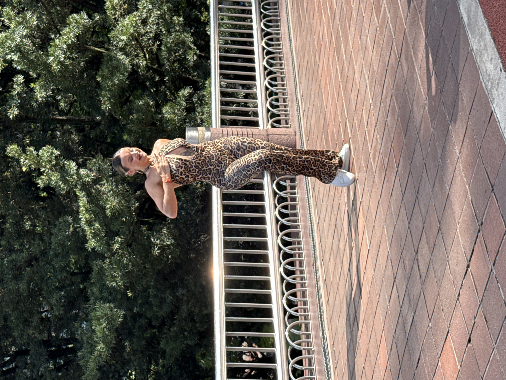 The photograph depicts a woman in a leopard-print jumpsuit standing on a brick-paved area adjacent to a metal railing. She is wearing white sneakers and has an orange wristband on her left wrist. The background features lush green trees. The location is Kuala Lumpur, Malaysia.