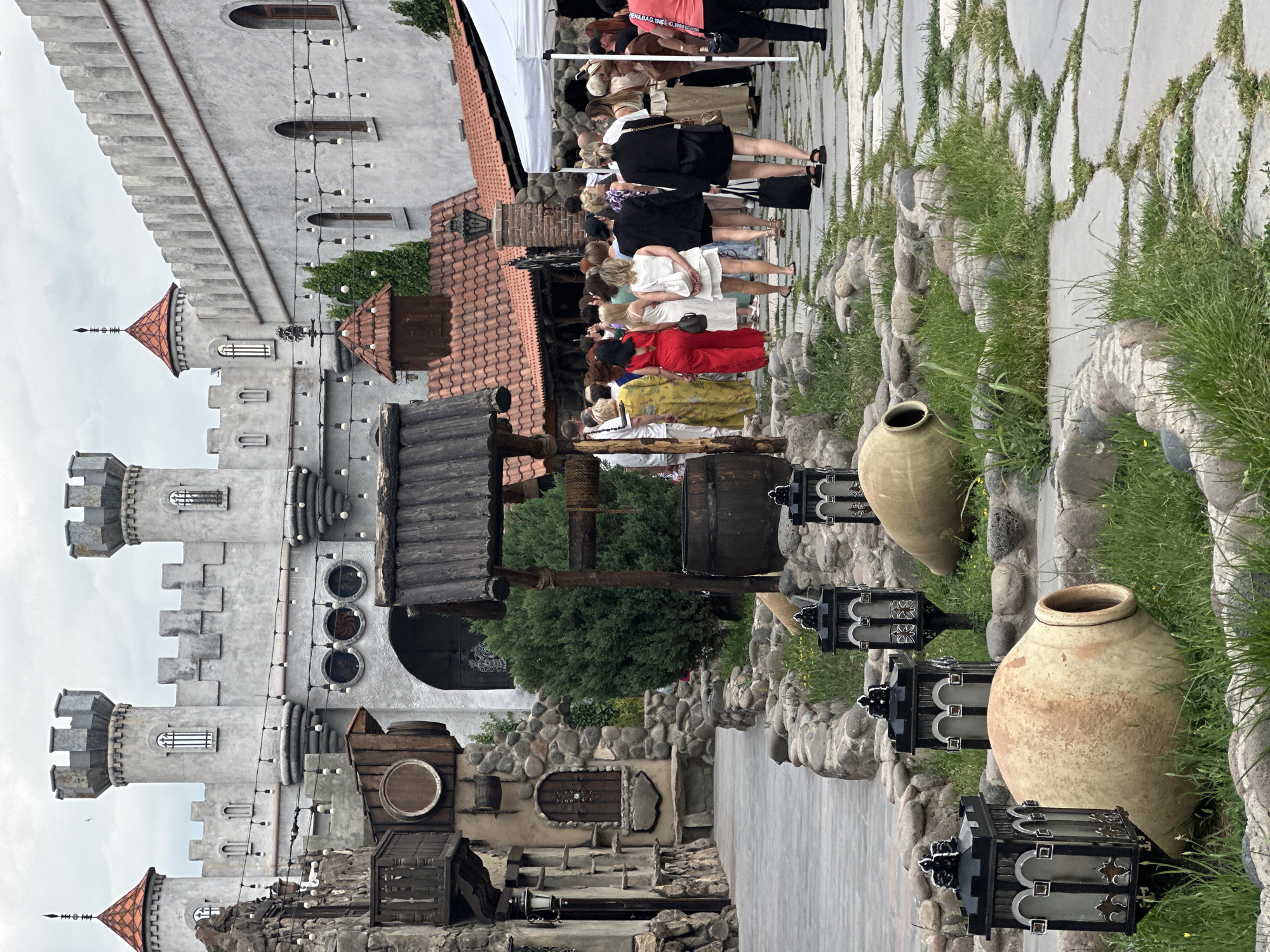 The image depicts a courtyard area, likely within a medieval-themed attraction or historical site in Voskevaz, Armenia. In the background, a large, stone-like castle structure dominates the scene with crenellated towers and arched windows, adorned with string lights. To the left, a rustic building with a wooden balcony and stone accents is visible. In the foreground, a cobblestone path winds through the courtyard, bordered by stone walls and lush green grass. Several large, earthenware pots and decorative lanterns are placed on the ground amidst the greenery. A wooden well with a barrel stands near the center of the image. A crowd of people, mostly women dressed in colorful and formal attire, are walking along the path, heading towards the right side of the frame, where a white tent structure is set up. The sky is overcast, suggesting a cloudy day.