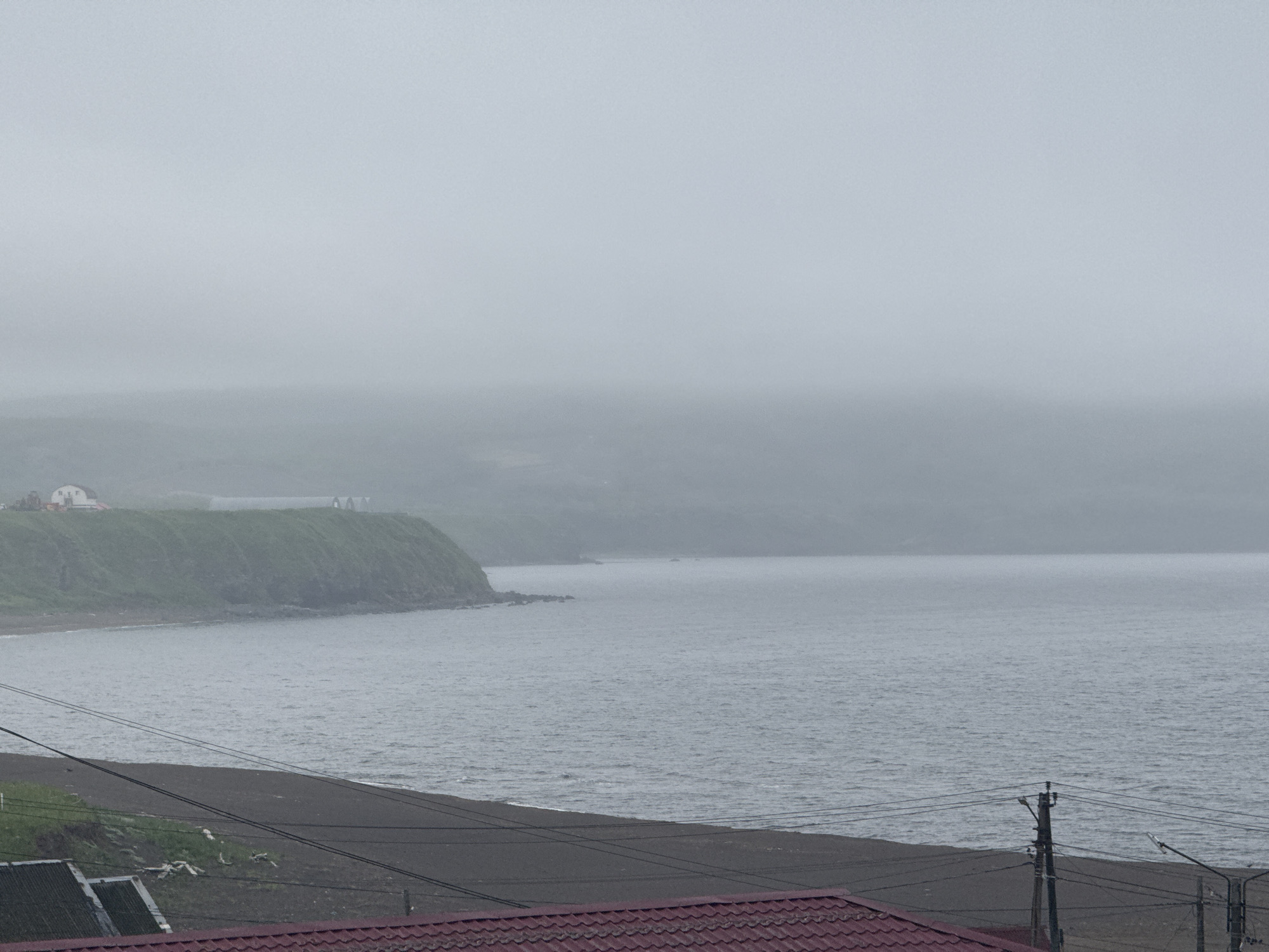 The image shows a view of the coast near Kuril'sk, Russia, under a heavy overcast sky. In the foreground, there is a dark-colored beach with some vegetation and debris. Several power lines are strung across the scene, supported by utility poles. Beyond the beach, the gray sea stretches towards a distant, fog-shrouded coastline. The land in the middle ground rises steeply, covered in green vegetation, with a few buildings visible on the top of the cliff. The overall atmosphere is somber and muted due to the weather conditions.