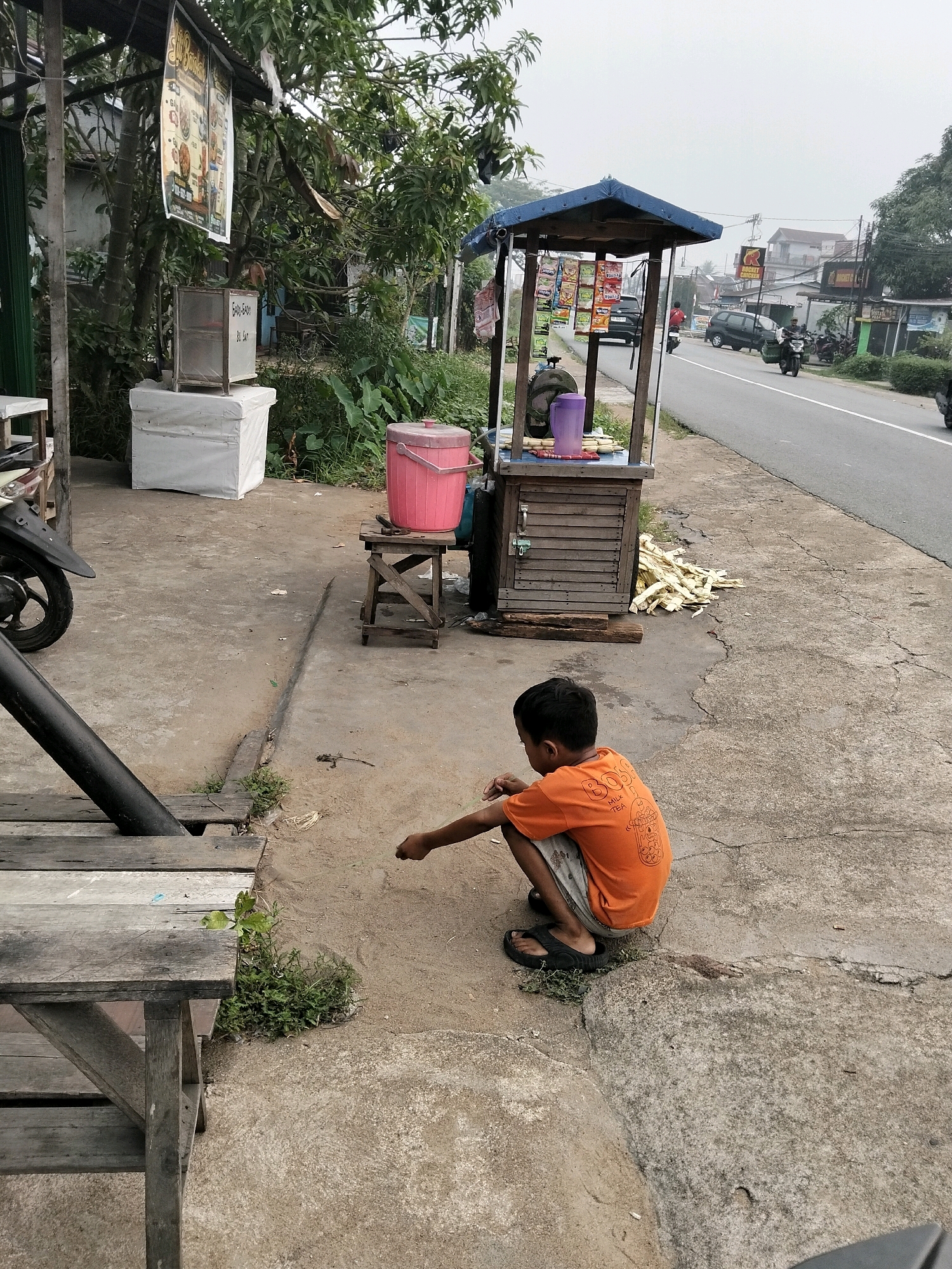 In Arang Limbung, Indonesia, a young boy wearing an orange t-shirt and shorts crouches on a sandy patch of ground next to a roadside stall. The stall, constructed of weathered wood and topped with a blue tarp, displays various packets of snacks. A pile of sugar cane stalks rests beside the stall, and a pink cooler sits on a wooden stool nearby. The boy appears to be drawing or playing in the dirt with his finger. In the background, a paved road stretches out, with cars and motorcycles moving along it. Buildings and lush green trees line the roadside, suggesting a semi-urban or rural setting. A wooden table and a portion of a motorcycle's wheel are visible in the foreground. The sky is overcast.