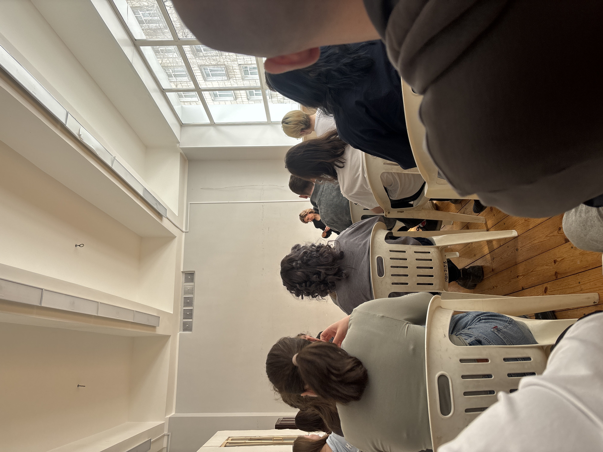 A classroom setting in Perm, Russia, shows a row of students sitting in white plastic chairs, seemingly focused on a lecture or presentation. The classroom is well-lit by natural light from a window. The students face forward, their backs visible to the viewer. The floor is wooden, and the room has white walls.