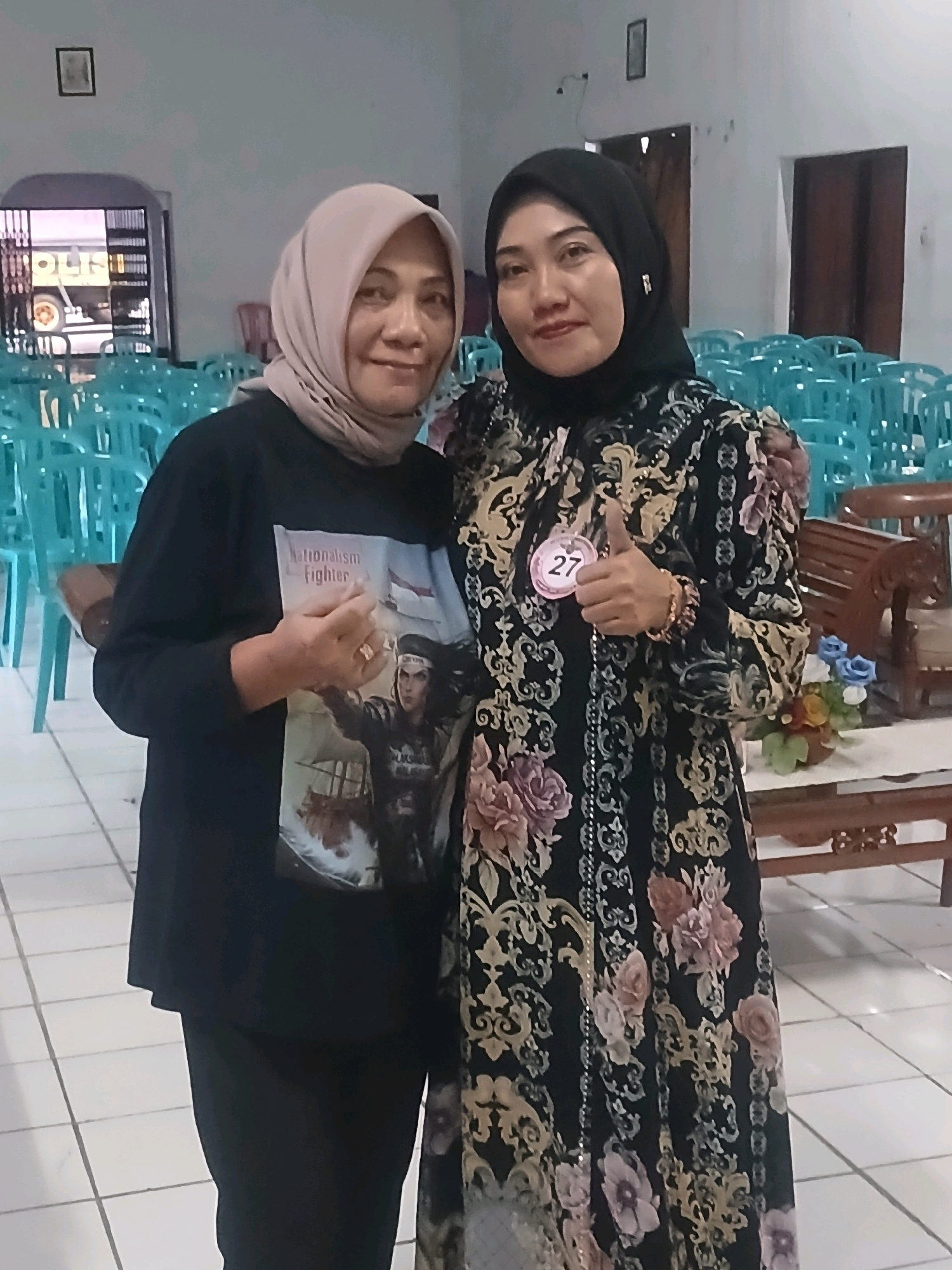 Two Indonesian women pose for a photo in what appears to be a community hall or meeting space in Banyuwangi, Indonesia. The woman on the left, wearing a light-colored hijab and a black long-sleeved shirt with a graphic print of a historical figure labeled "Nationalism Fighter," smiles at the camera. The woman on the right, wearing a black hijab and a black and gold floral patterned dress, smiles and gives a thumbs-up. She wears a circular pink badge with the number "27" on her dress. The background shows rows of teal plastic chairs, typical of event venues in Indonesia, and a table with a vase of artificial flowers. The floor is tiled with a white and light gray grid pattern. A glimpse of a yellow truck is visible through a window in the background.