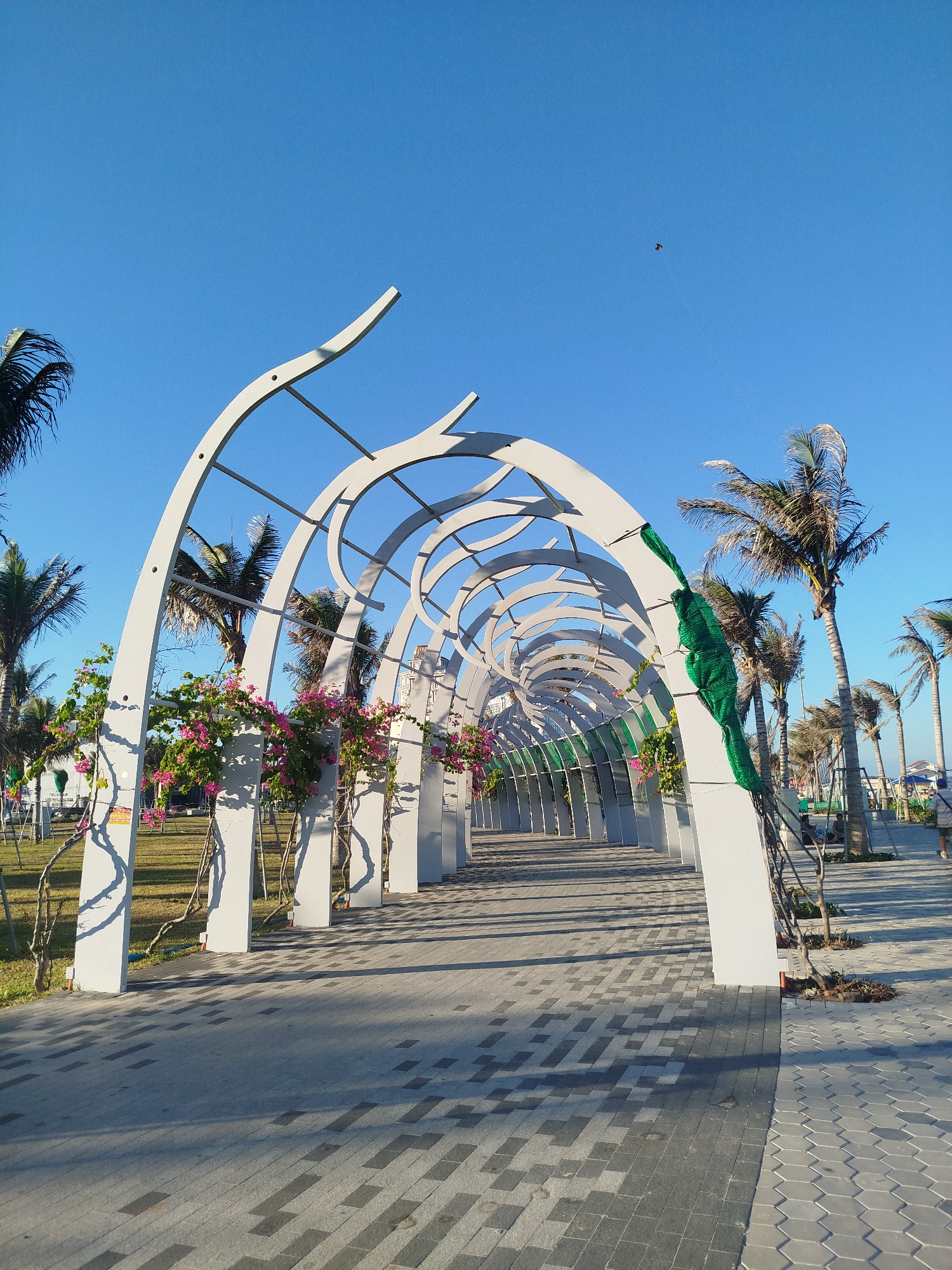 The image depicts an outdoor pathway lined with a series of white, arched structures, creating a tunnel-like effect. The pathway is paved with gray and black interlocking blocks. Between the structures are rows of palm trees and small patches of green grass. Some of the arches are decorated with pink bougainvillea flowers, and green netting is draped over one of the arches. A person walks along the pathway in the distance. The sky above is clear and blue. A kite is visible in the distant sky. The location is likely a park or public space in Ho Chi Minh City, Vietnam.