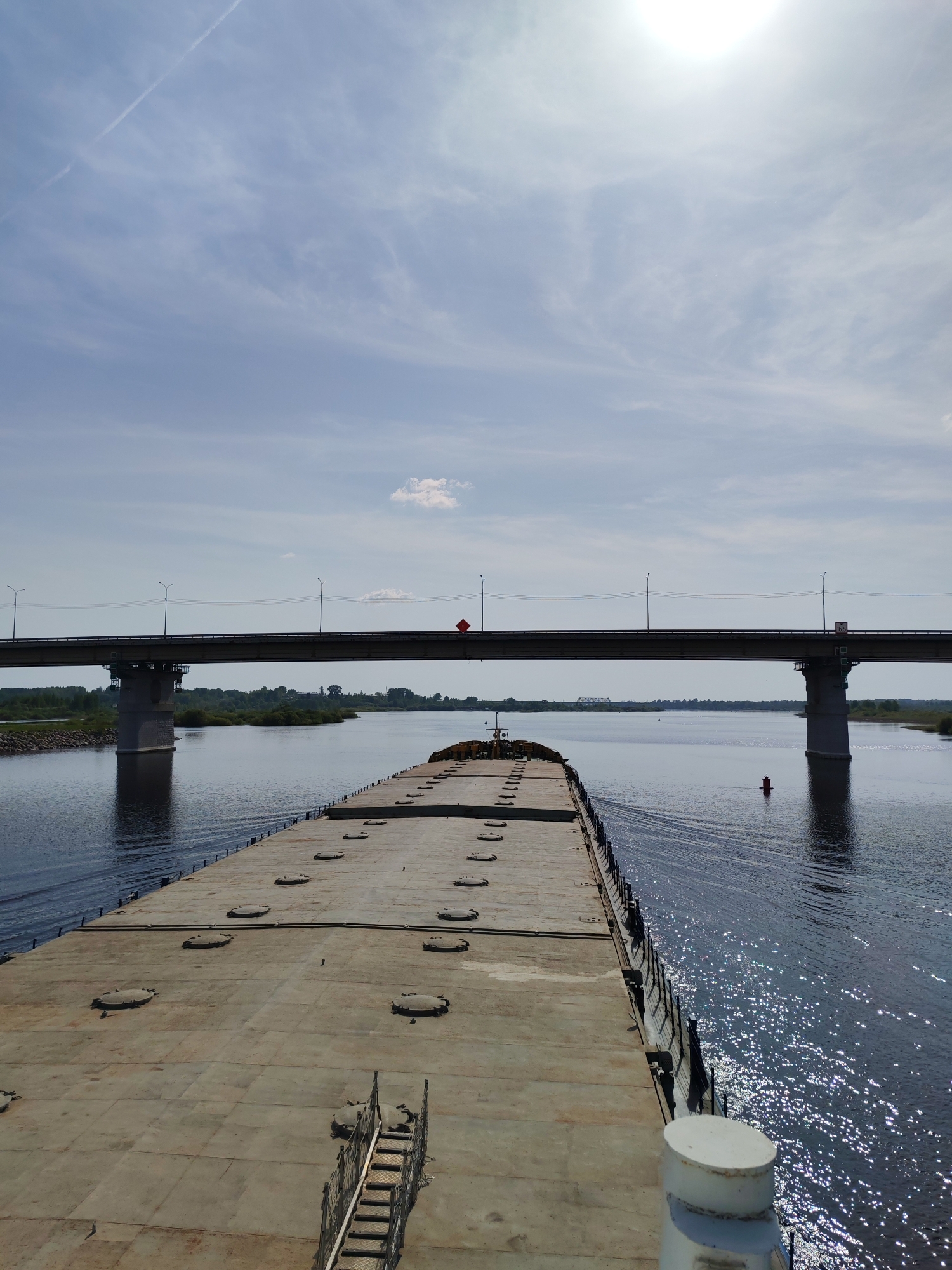 The image depicts a large, flat-decked barge navigating a wide river under a concrete bridge in the settlement of Нифантово, Russia. The barge occupies a significant portion of the foreground, with numerous circular hatches visible on its deck. A metal staircase is present on the deck. The bridge, supported by substantial pillars, spans the river in the middle distance, with foliage and vegetation visible along the riverbanks. A red buoy is visible in the water. The sky is pale blue with scattered clouds and the sun is bright and partially obscured by cloud cover. The river's surface is slightly rippled and reflects sunlight.