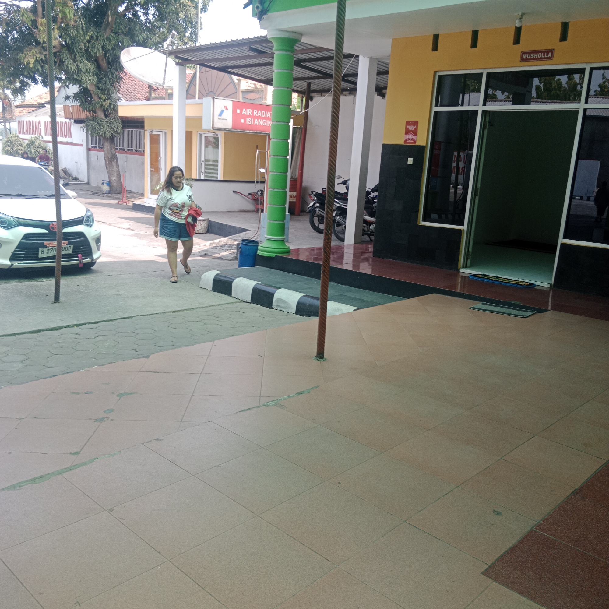 The image depicts an outdoor scene at what appears to be a gas station or service area in Taraman, Indonesia. In the foreground, the ground is paved with light brown and grey square tiles, some of which show signs of wear and staining. A black and white striped speed bump is visible on the left side. To the left of the speed bump, a white car is parked.
A woman wearing a light-colored t-shirt with a graphic and denim shorts walks towards the right of the frame, carrying a red garment over her arm. She is wearing sandals. Behind her, a white car is visible, with a license plate that reads "B 270 UOB".
In the background, a building with a yellow facade and a dark grey base is present. A sign above the entrance reads "MUSHOLLA", indicating a prayer room. To the left of the building, a white structure with an awning and a satellite dish is visible. There are signs on the building, one red and white reads "AIR RADIATOR ISI ANGIN". Further to the left, a red and white sign with a crossed-out cigarette symbol reads "DILARANG MEROKOK" (No Smoking). Several motorcycles are parked under the awning of the building. A blue bin is situated near a green pillar. The overall environment suggests a public space with facilities for services and amenities.