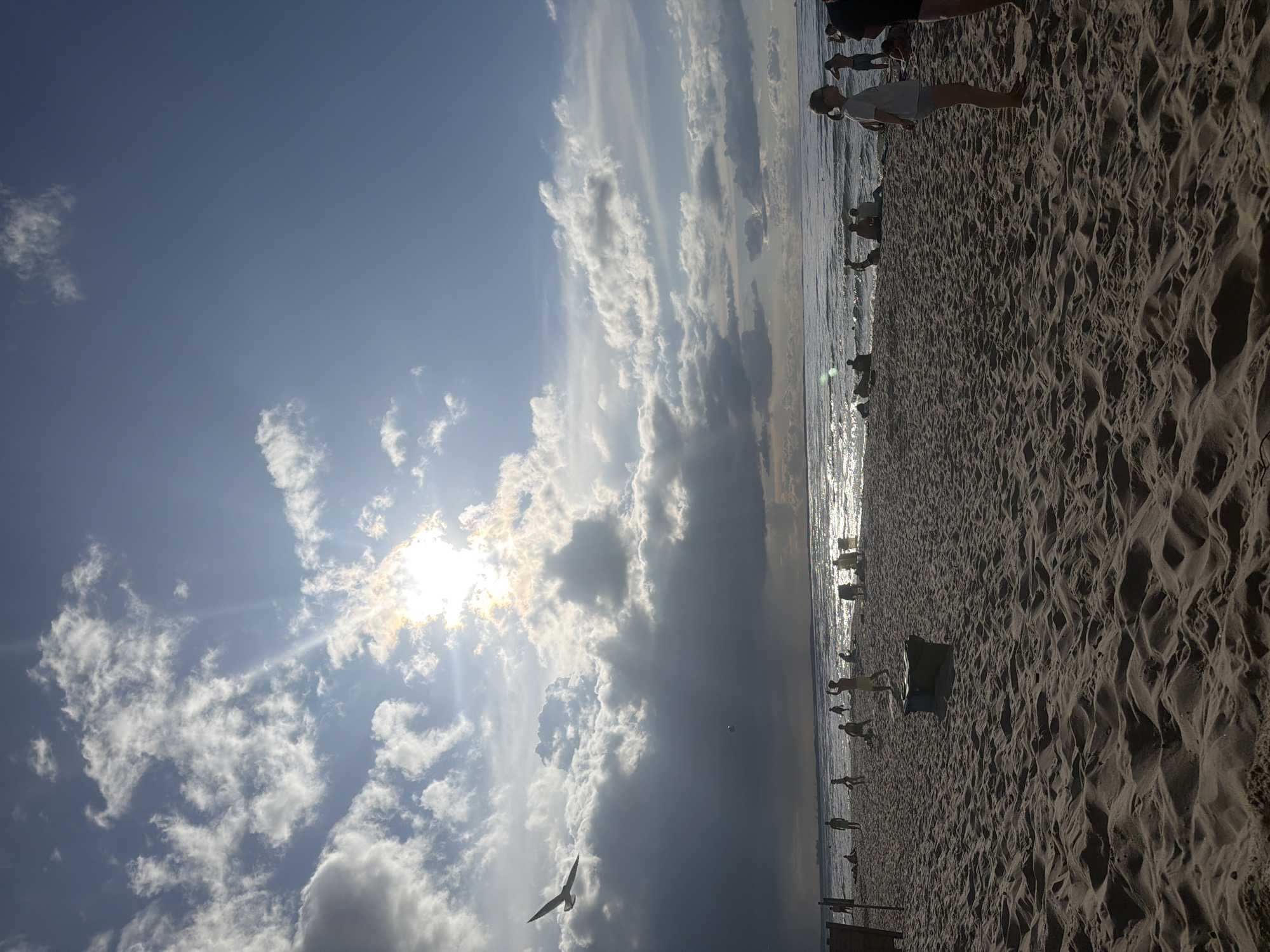 A wide-angle, full-frame shot captures a beach scene in Międzyzdroje, Poland, under a dramatic sky. The sun, partially obscured by clouds, casts brilliant rays across the water and illuminates the sky with a bright, ethereal glow. The foreground is dominated by textured sand dunes, leading to a beach populated by various individuals. Some are engaged in activities, like playing, while others are seated or standing near the water's edge. A few people are visible in the water, interacting with the waves. In the distance, the ocean stretches out, its surface shimmering with sunlight. A bird is in flight against the backdrop of the sky and clouds. The overall atmosphere is one of leisure and natural beauty, with the dynamic sky adding a sense of drama to the scene.