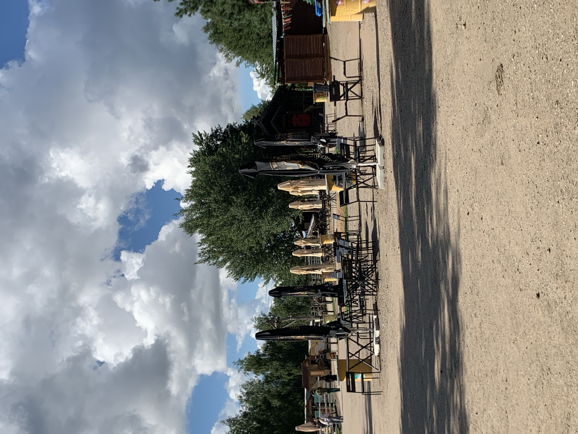 An outdoor cafe area is depicted in this image, set against a backdrop of lush green trees and a partly cloudy sky. The ground is covered in a sandy, gravelly surface, and the scene is sparsely populated. Several tables and chairs are arranged, some with large black or beige umbrellas folded down, suggesting they are not in use. A few people are visible in the background, walking near some structures that appear to be small buildings or stalls. The overall impression is one of a quiet, possibly early-season or off-peak moment at a relaxed, open-air venue. The image is likely taken in Baranavichy, Belarus, given the context.