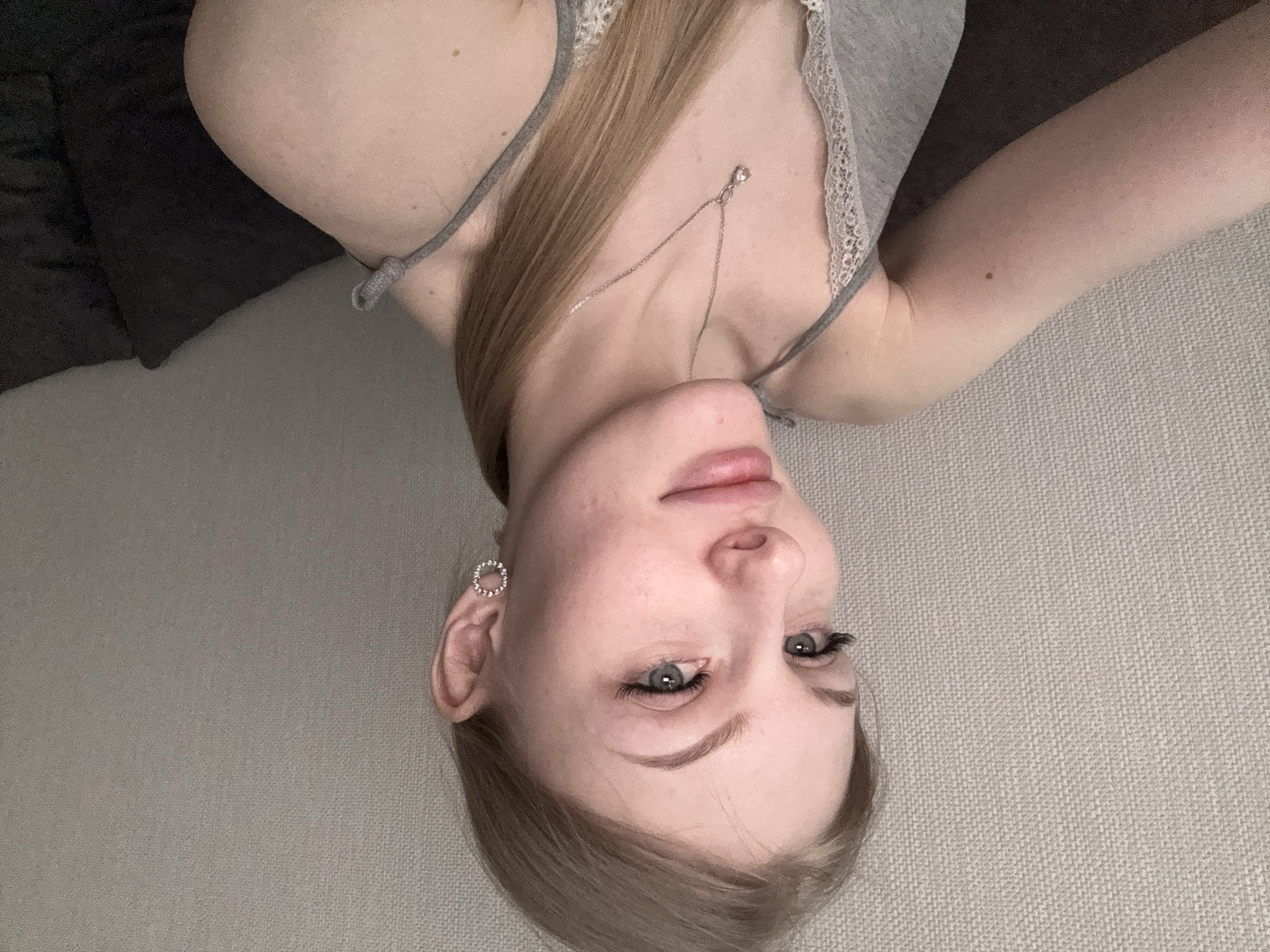 An indoor portrait image depicts a young adult female in Perm, Russia, looking directly towards the camera with a subtle smile. She possesses light skin, light brown hair styled in a low ponytail visible on her right side, and blue-grey eyes with pronounced eyelashes. Her eyebrows are light brown and well-defined. She is dressed in a grey camisole featuring white lace trim along the neckline and thin straps. A silver-colored chain necklace with a small, clear teardrop-shaped pendant is visible around her neck. Her left ear is adorned with a circular stud earring set with small stones. A distinct dark brown mole is present on her right upper arm. The background consists of a light grey textured wall, and a dark, indistinct object, likely a cushion, is partially visible to her right. The scene is evenly lit, indicating an interior setting.