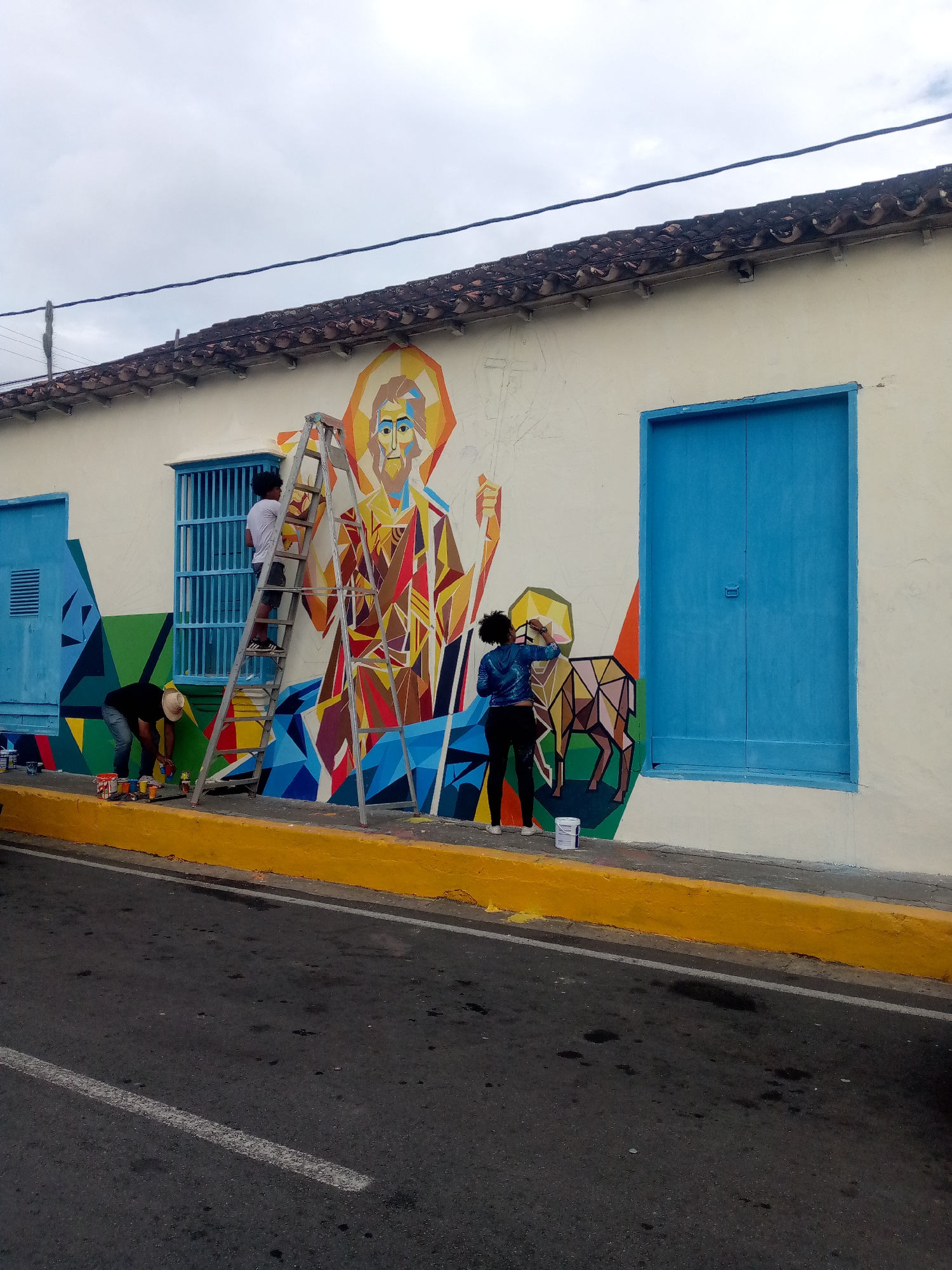 In San Juan Bautista, Venezuela, two individuals are actively engaged in painting a vibrant mural on the exterior wall of a building. The mural depicts a stylized figure with a halo, rendered in geometric shapes and a bright color palette, and an animal figure also composed of geometric facets. A ladder is positioned centrally, allowing one of the painters, a person wearing a white t-shirt and dark shorts, to reach higher sections of the artwork. Another painter, dressed in a black t-shirt and a straw hat, is on the ground, tending to paint cans and smaller brushes. A third individual, wearing a blue jacket and black leggings, is also on the ground, meticulously painting a section of the mural. The wall itself is painted a neutral off-white, contrasting with the colorful mural. A prominent, solid blue double door is visible to the right of the mural. A bright yellow painted curb runs along the base of the wall, separating the sidewalk from the asphalt street. The sky above is overcast.