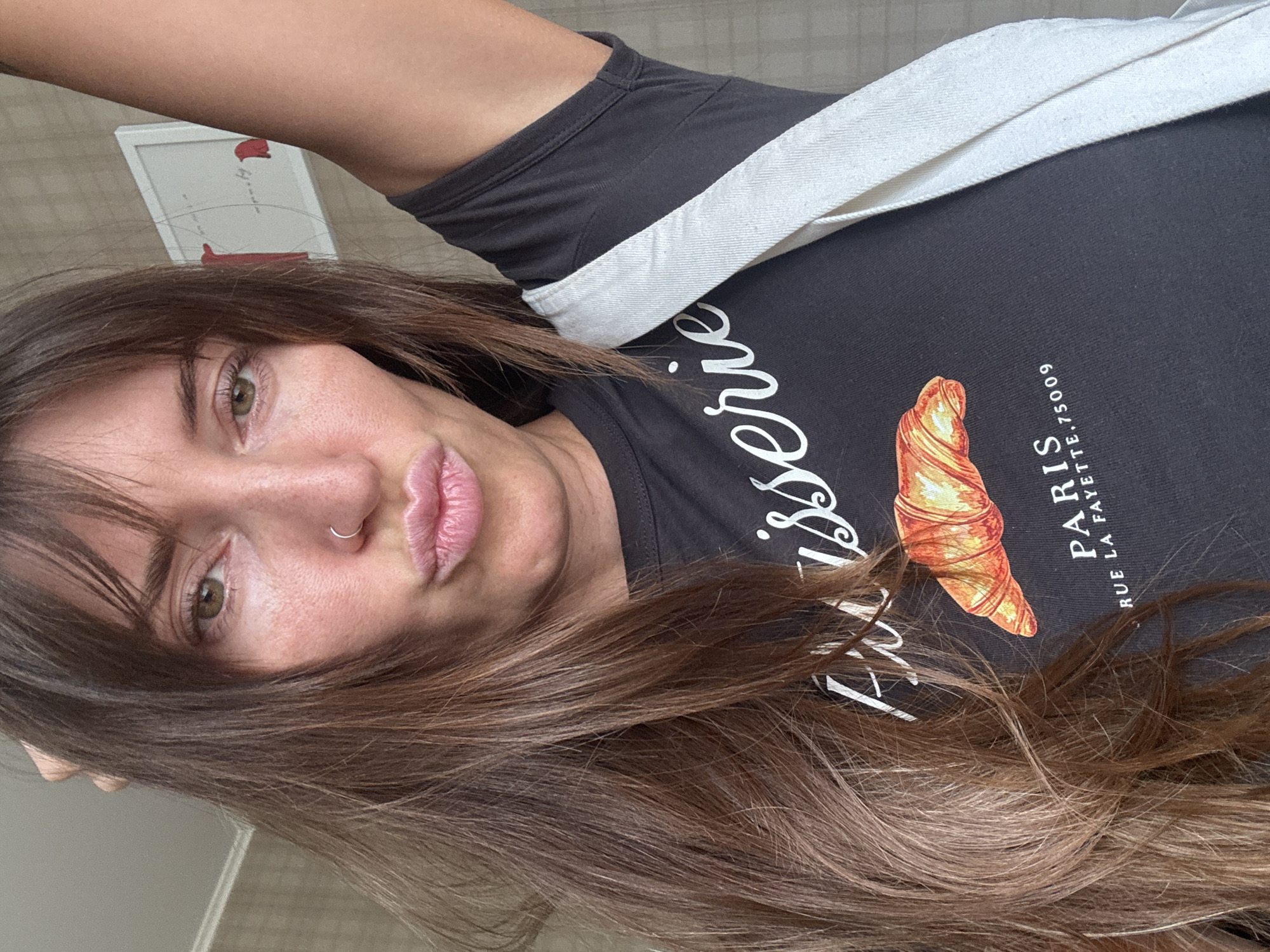 A woman with long brown hair, wearing a dark t-shirt with a croissant and "Paris Rue La Fayette, 75009" printed on it, poses for a selfie. She has a nose ring and her lips are pursed. Her right arm is raised and a white strap from a bag is visible across her chest. The background appears to be an indoor setting with a light-colored wall and a framed picture. There are no visible indications of the image being taken in Moscow, Russia.