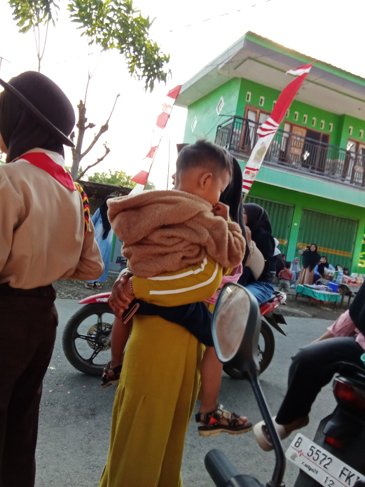 The image captures a street scene in Bungatan, Indonesia, likely during an outdoor event or gathering, given the presence of Indonesian flags. The foreground is dominated by people and a motorcycle. On the left, a person wearing a brown uniform with a red scarf, reminiscent of scout attire, is partially visible from the back. In the center, an adult, dressed in a long yellow garment, is carrying a toddler who is wrapped in a brown blanket. The toddler is facing away from the camera, looking towards a green building in the background. The building has a balcony and several windows, adorned with more red and white flags. Behind the central figures, other people are visible, some sitting at tables and others standing. Another motorcycle is parked in the foreground on the right, with its license plate showing "B 5572 FK". The ground is paved asphalt. The overall lighting suggests it is late afternoon or early evening, with the sun casting a warm glow.