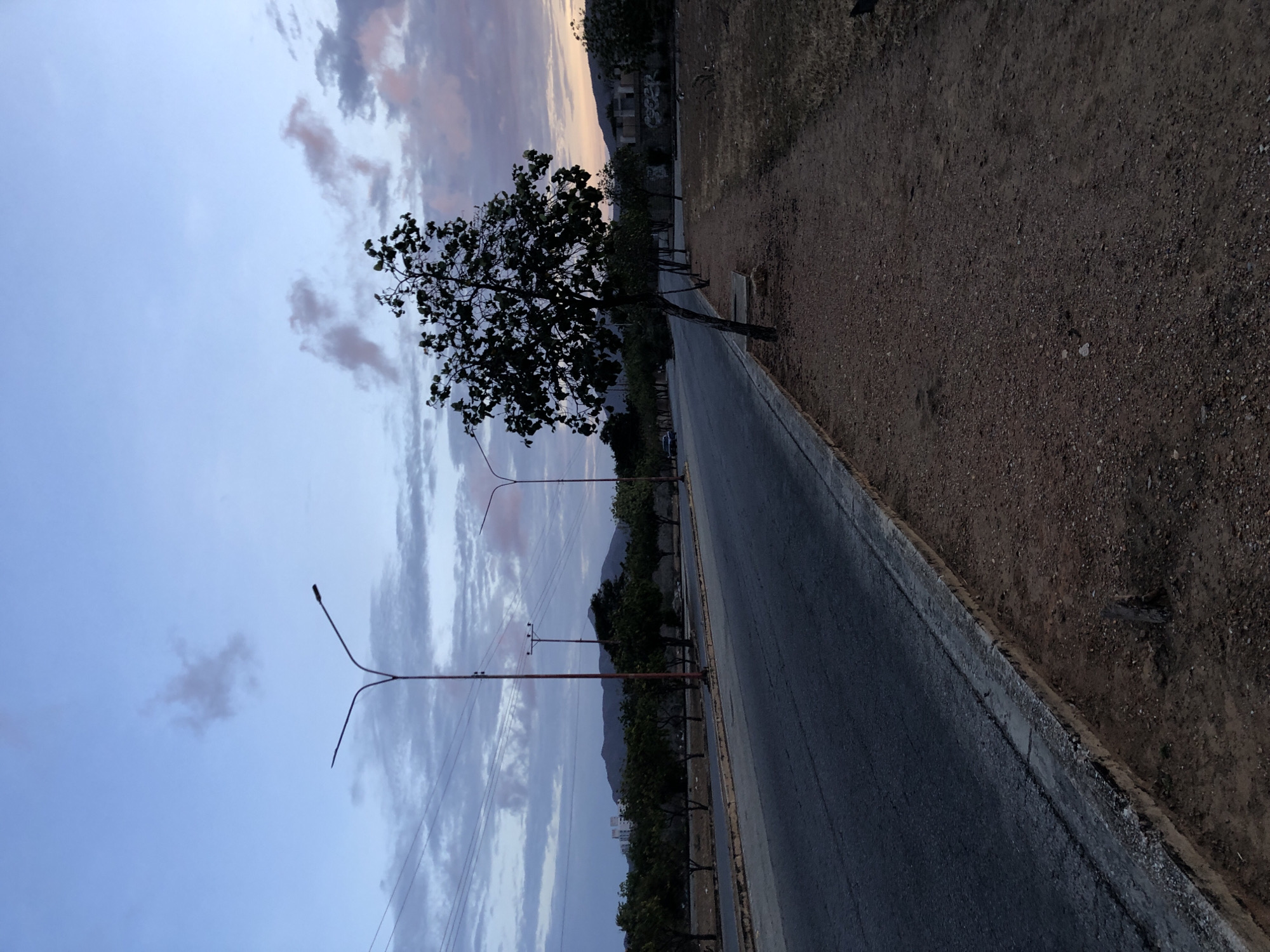 The image depicts a road with a tree on the left side and a dirt shoulder on the right. There are streetlights along the road, and in the distance, there is a mountain range. The sky is cloudy with a hint of sunset colors. Based on the urban landscape, the presence of mountains, and the provided location, it is likely somewhere within the outskirts or less densely populated areas of Juan Griego, Venezuela. There are no people visible in the scene. The road seems to be a secondary road or access road, rather than a major highway. The buildings in the background seem to be residential.