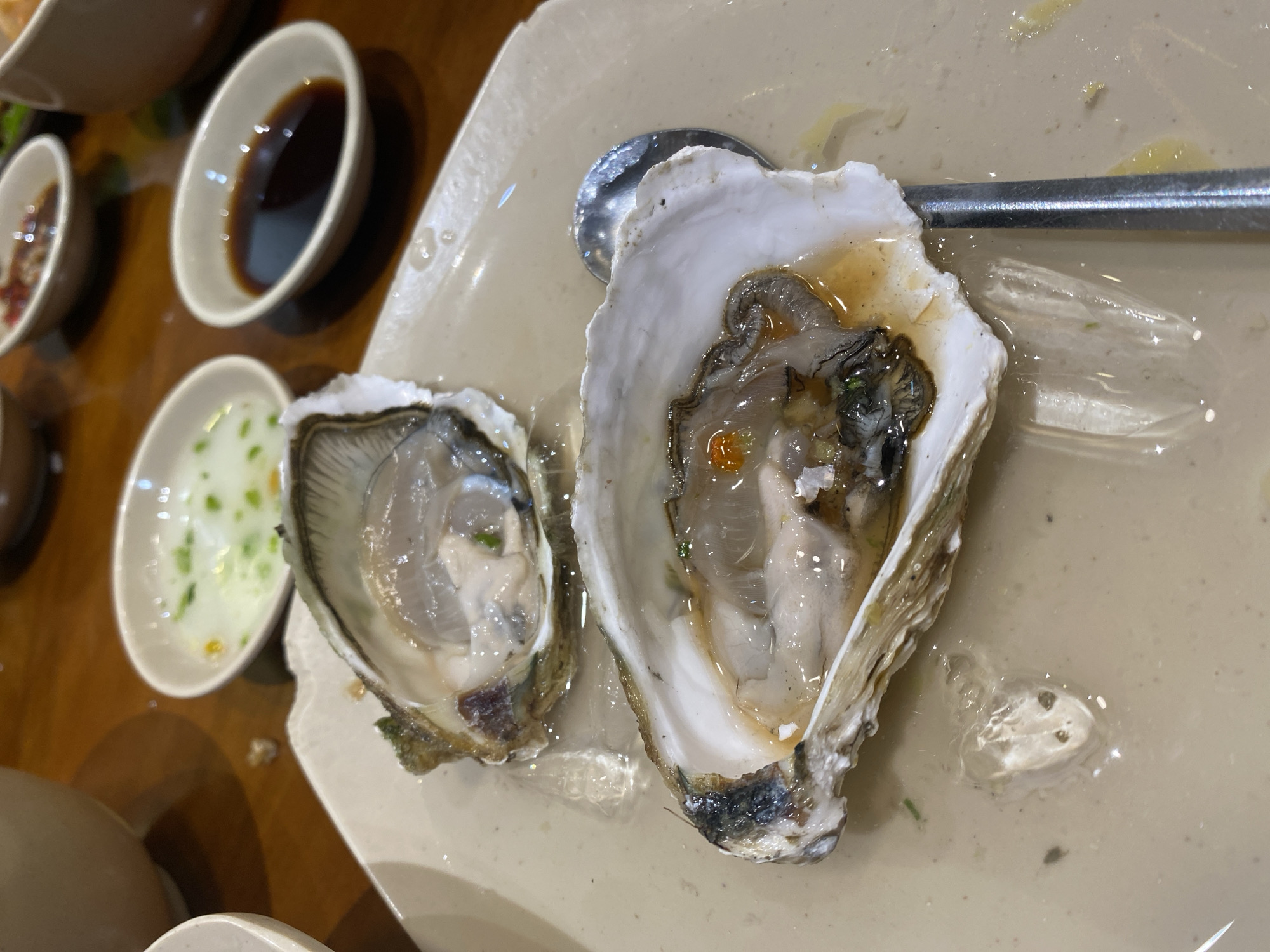 This is a close-up, top-down shot of two opened oysters on a light-colored plate. The oysters are served in their shells, with the fleshy part visible and glistening. One oyster is larger and positioned to the right, while a smaller one is to the left. Both appear to be raw and served with some liquid, possibly their natural juices or a light sauce. A metallic spoon rests partially on the larger oyster and extends towards the right edge of the frame.
In the background, to the left of the plate, are several small white dipping bowls. One contains a dark liquid, likely soy sauce or a similar condiment. Another contains a lighter, creamy sauce with visible green specks, possibly chopped herbs or scallions. The table surface appears to be a warm-toned wood. The lighting suggests an indoor setting, possibly a restaurant.
The image captures the presentation of a seafood dish, likely an appetizer. The focus is on the oysters and their accompaniments, suggesting a meal in progress or about to be consumed. The overall impression is one of freshness and culinary preparation. No people are visible, and the scene is focused solely on the food and serving ware. The time of day and weather are not discernible from the image. No visible text is present. The location context provided is Nha Trang Ward, Vietnam, which is consistent with a coastal region known for its seafood.