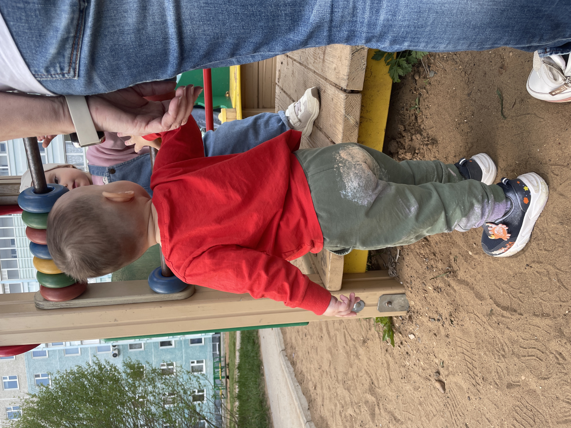 The image shows a young child in a red long-sleeved shirt and green pants with what appears to be grass stains on the knees, standing on what looks like a wooden playground structure in Perm, Russia. The child is wearing black and white sneakers and is reaching for a metal fastener on the playground. Another child wearing a pink shirt and a woman's arm with a smartwatch are visible to the left. Above them is a person wearing blue jeans. The ground is sandy. In the background, there is a multi-story building. The sky is not visible.