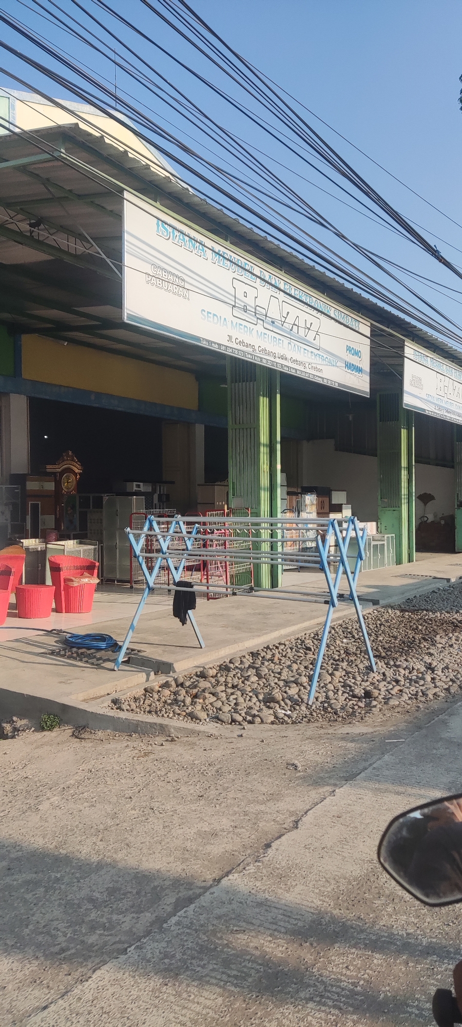 A wide shot captures the exterior of a commercial establishment, likely a furniture and electronics store, in Gebang, Indonesia. A large banner hangs across the front, bearing the text "ISTANA MEUBEL DAN ELEKTRONIK" and additional information about branches and contact details. The store's facade features a green metal gate system and displays various furniture items and appliances inside. In the foreground, a blue metal drying rack with a black piece of clothing hanging on it is positioned on a concrete pavement. A coiled blue hose is also visible on the ground near the drying rack. The area surrounding the store appears to be an open space with a gravel-covered section. Power lines crisscross the sky above, indicating an urban or suburban setting. The overall scene is well-lit by natural sunlight, suggesting it is daytime.