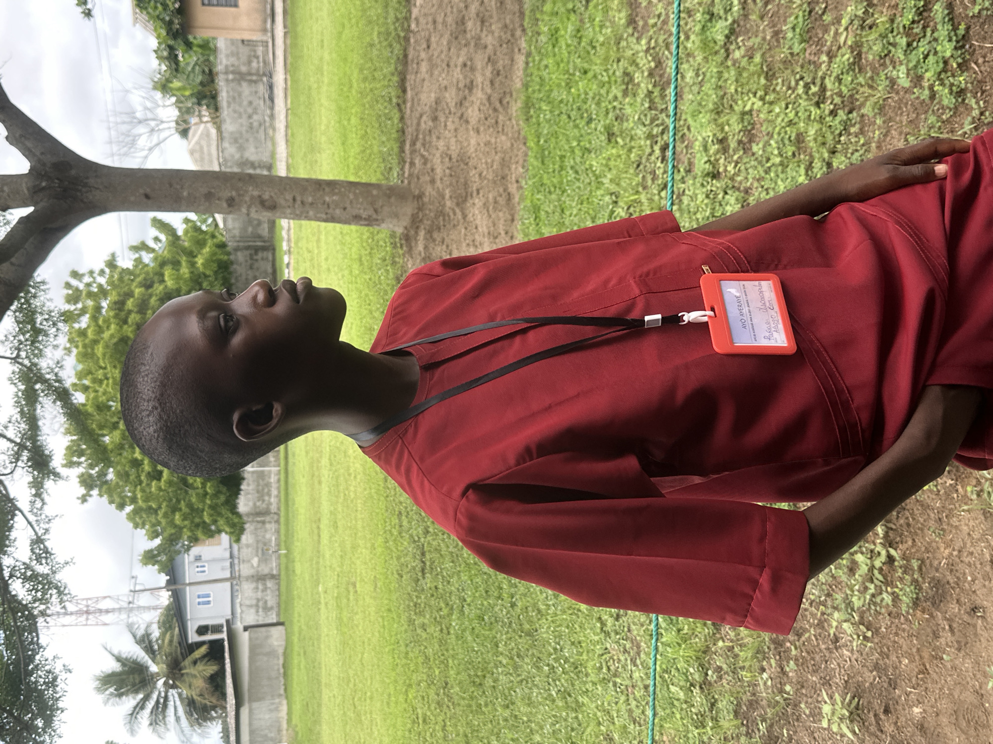 A young Black individual, appearing to be male, stands outdoors wearing a dark red, short-sleeved tunic with a high neckline and subtle decorative stitching on the chest. A black lanyard with a white clip is around his neck, holding a bright orange identification card with text and handwriting. His left hand is tucked into the pocket of his tunic. He has a short buzz cut and is looking to his right with a neutral expression. The background consists of a lush green lawn with scattered patches of dry earth and a thin green rope fence running horizontally across the lower portion of the frame. A large tree trunk is visible to the right of the individual. In the distance, beyond the lawn, there are buildings, including one with white trim and blue windows, and a tall radio tower. The sky is overcast. The overall setting appears to be a school or community grounds in Itoga, Nigeria.