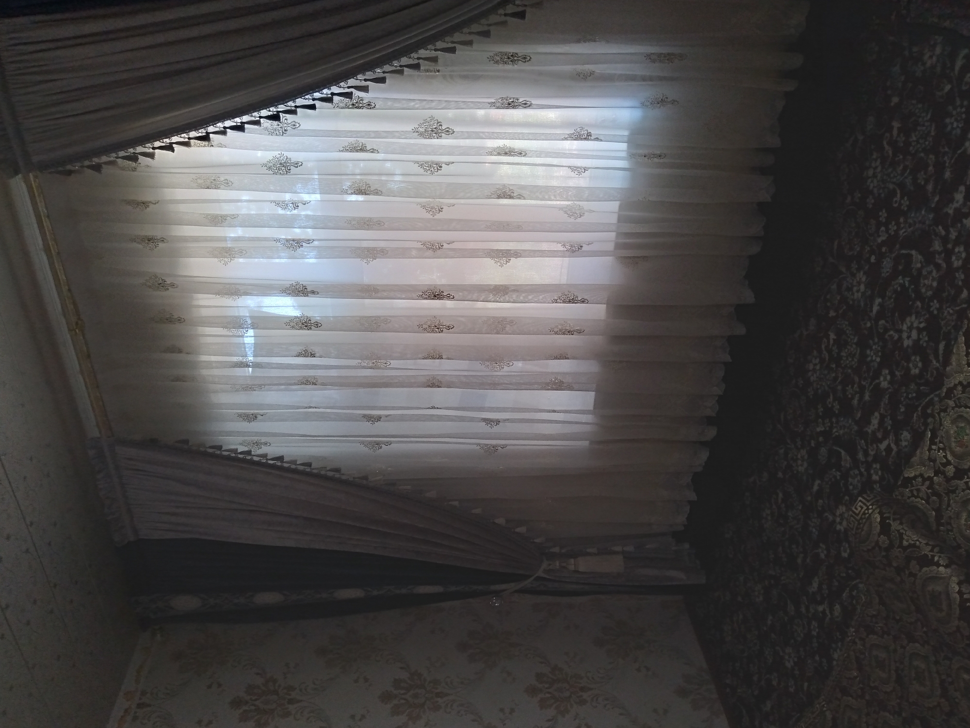 A dimly lit, interior shot of a room with patterned wallpaper and a dark, floral carpet. A large window is covered by sheer, white curtains adorned with a repeating damask-like pattern. Two sets of heavier, taupe-colored curtains are drawn to the sides, one on the left and one on the right, each decorated with a fringe of dark tassels. A thick, taupe tie-back with a decorative tassel secures the left curtain. The ceiling is textured and appears to be a light color. The overall impression is of a residential interior, likely a living room or bedroom, in the city of Baki, Uzbekistan.