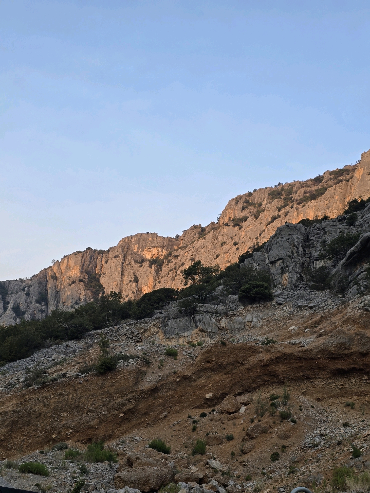 The image depicts a mountainous landscape under a clear, pale blue sky. The foreground is a rocky, uneven slope covered with sparse vegetation, including small bushes and rocks of various sizes. The middle ground features a steeper, rugged cliff face, with some sections illuminated by warm, golden sunlight, suggesting it is either sunrise or sunset. Dark green trees and shrubs are scattered across the lower parts of the cliff face and the slope. The upper part of the image is dominated by the sky, with a very subtle gradient of color. The overall impression is one of a natural, rugged terrain.
Based on the landscape and vegetation, this scene is likely located in the mountainous region surrounding Podgorica, Montenegro. Specifically, the terrain, rock formations, and the type of flora visible are consistent with the karst landscapes found in the Dinaric Alps, which are prevalent in Montenegro and near the capital city.