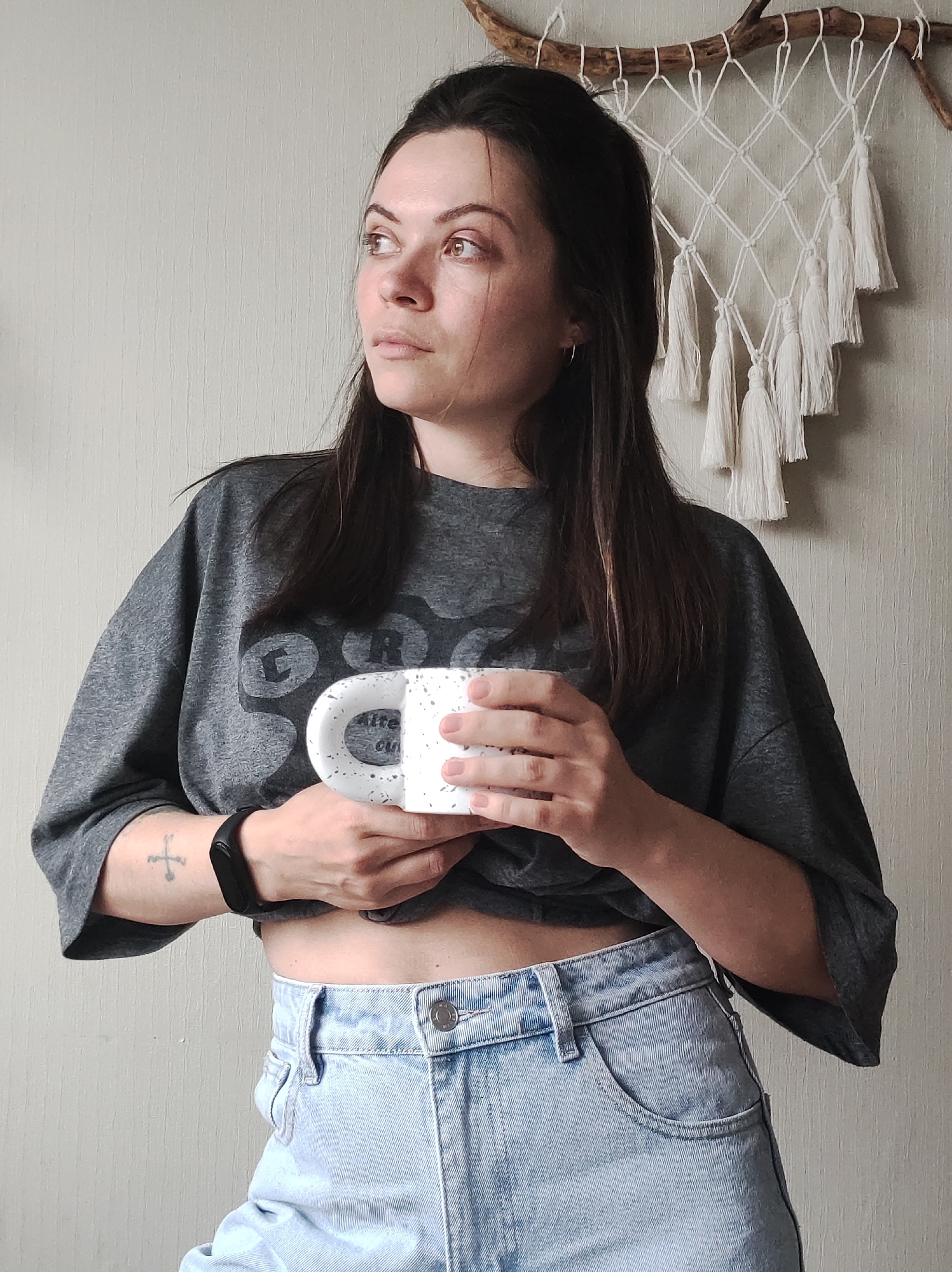 A young woman with dark hair and a gray t-shirt holds a white speckled mug, standing indoors against a light-colored wall. The t-shirt is cropped to expose some of her abdomen above light-wash jeans. She wears a black fitness tracker on her left wrist, below a cross tattoo. Behind her, a macrame wall hanging is suspended from a branch. No evidence suggests this photo was taken in Chaikovsky, Russia.