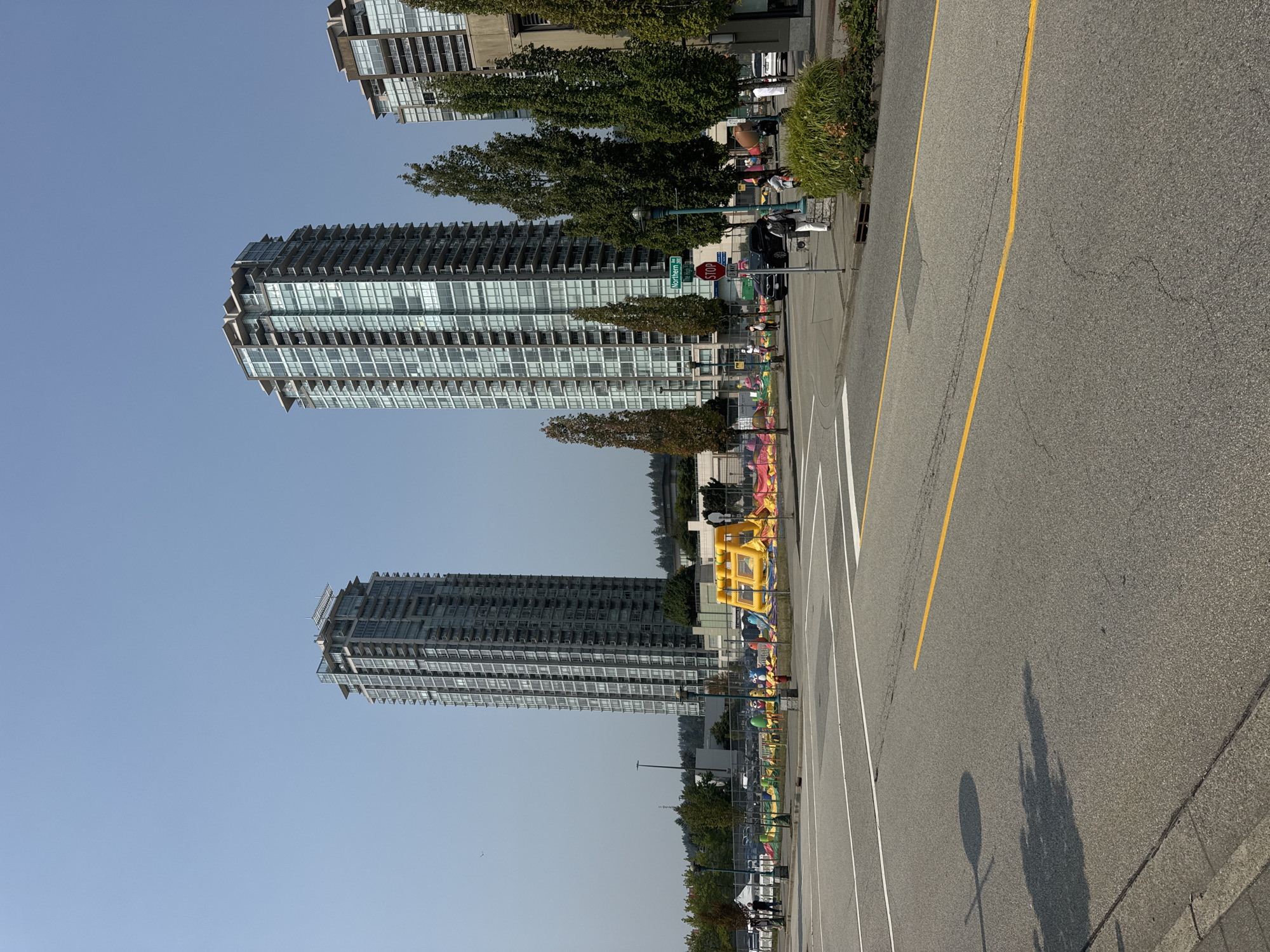 The image shows an outdoor scene with two prominent high-rise apartment buildings in the background. In the middle ground, there is a temporary inflatable playground with various colorful structures, including a large yellow inflatable slide. A chain-link fence encloses the playground area. Several people are visible walking around near the playground and on the sidewalk. A street sign indicating "Northern Rd" and "3000" is visible. Several trees are planted along the sidewalk. In the foreground, there is an asphalt road with clear lane markings, including a solid yellow line. The sky is clear and blue. The location is the intersection of Northern Road and Barnet Highway in Coquitlam, Canada.