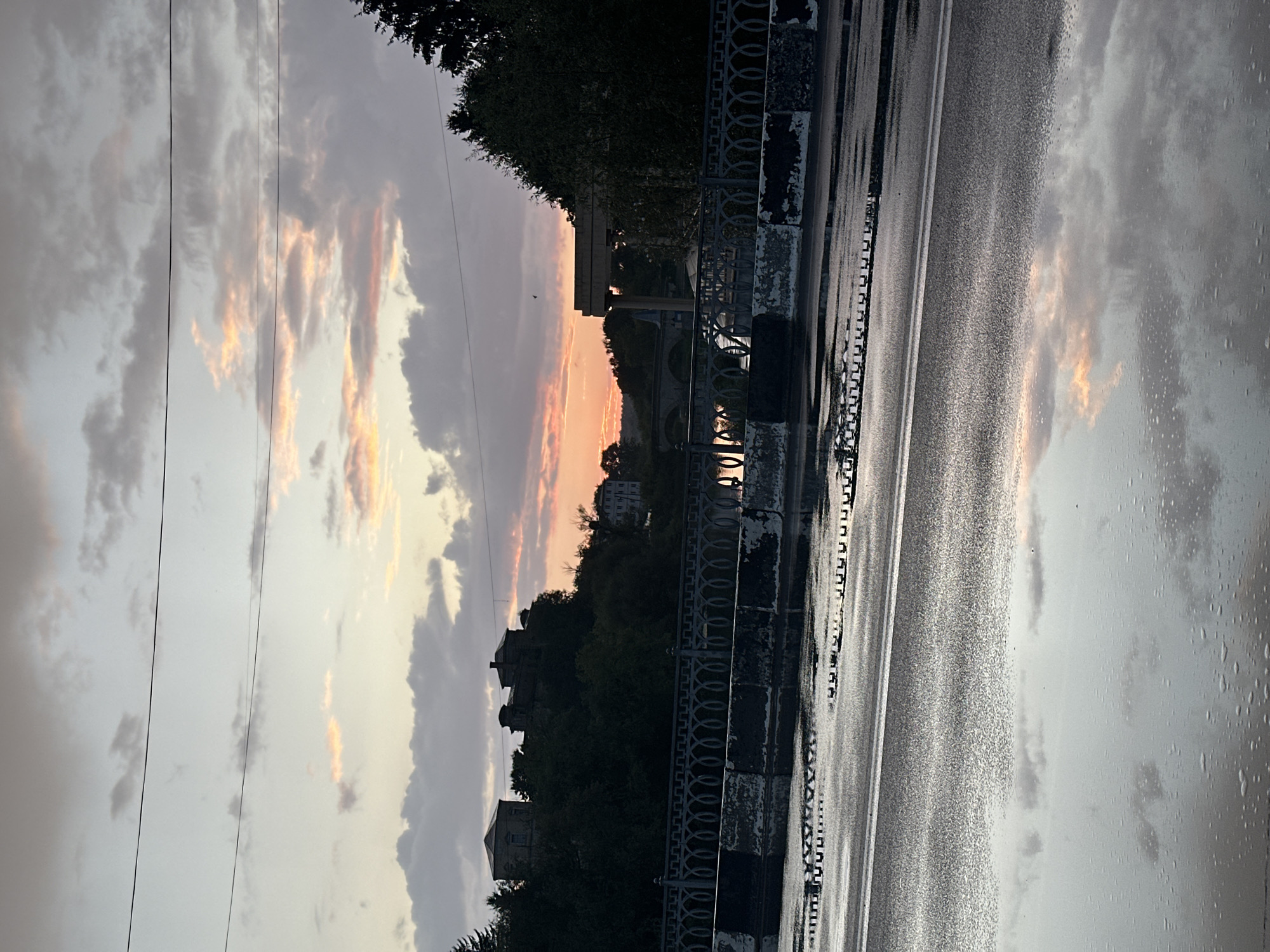 The photograph captures a twilight scene in Khmilnyk, Ukraine. The foreground shows a wet asphalt road, with large puddles reflecting the sky and surrounding environment. A section of a guardrail, constructed from metal and concrete barriers, runs across the middle ground. Beyond the guardrail, a body of water, possibly a river, is visible. The background is dominated by a hill covered in dense trees, with several buildings silhouetted against the sky on the left side. On the right, trees and an architectural structure are visible. The sky is filled with clouds tinged with the warm colors of sunset, with power lines stretching horizontally across the upper portion of the frame. The overall impression is one of a tranquil evening in a Ukrainian town.
