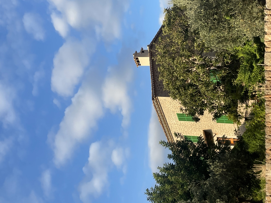 A traditional stone house is depicted from a low angle under a blue sky with scattered white clouds. The structure, located in Deià, Spain, features a light brown, rough-hewn stone facade. Multiple rectangular windows, each fitted with closed green louvered shutters, are visible on at least two stories. A terracotta tiled roof caps the building, from which a distinctive, multi-tiered, light-colored chimney rises on the right side. Dense green foliage, including trees with varying leaf densities and what appears to be an olive tree on the right, partially obscures the house. A reddish-brown door is partially visible on the left side of the house. In the immediate foreground, a dry-stone wall constructed of light-colored, irregular stones spans the width of the frame. No individuals, animals, or active interactions are present in the scene. The environment is well-lit by natural sunlight.